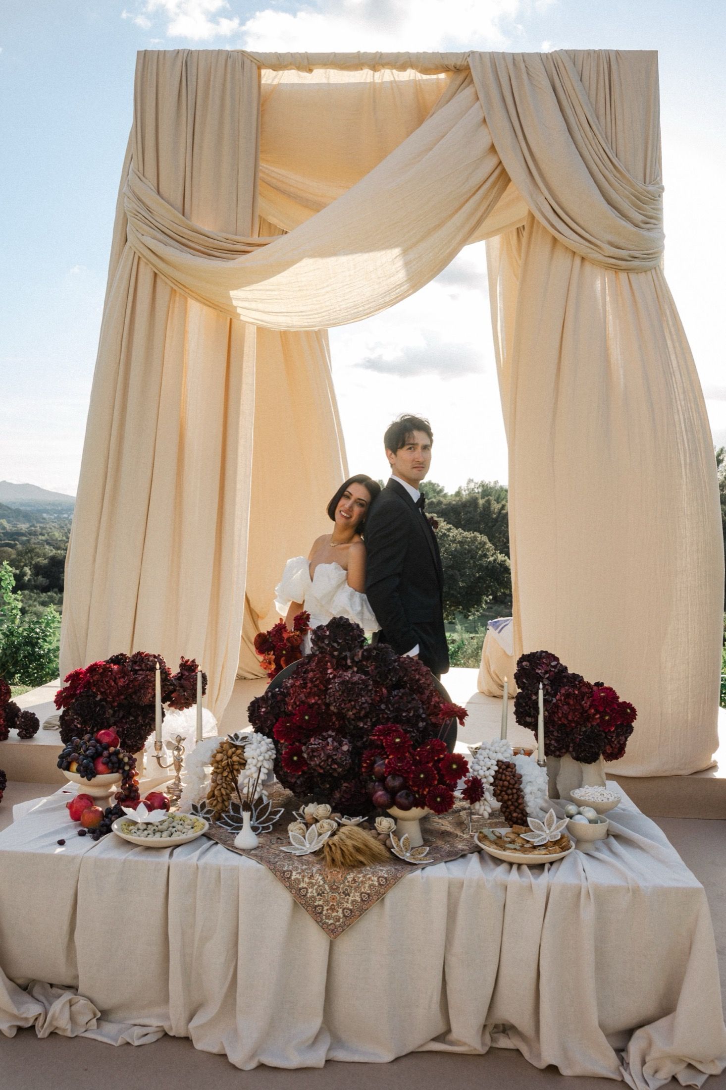 Elegant wedding table setup with deep red floral arrangements and draped cream fabric canopy at an outdoor venue.