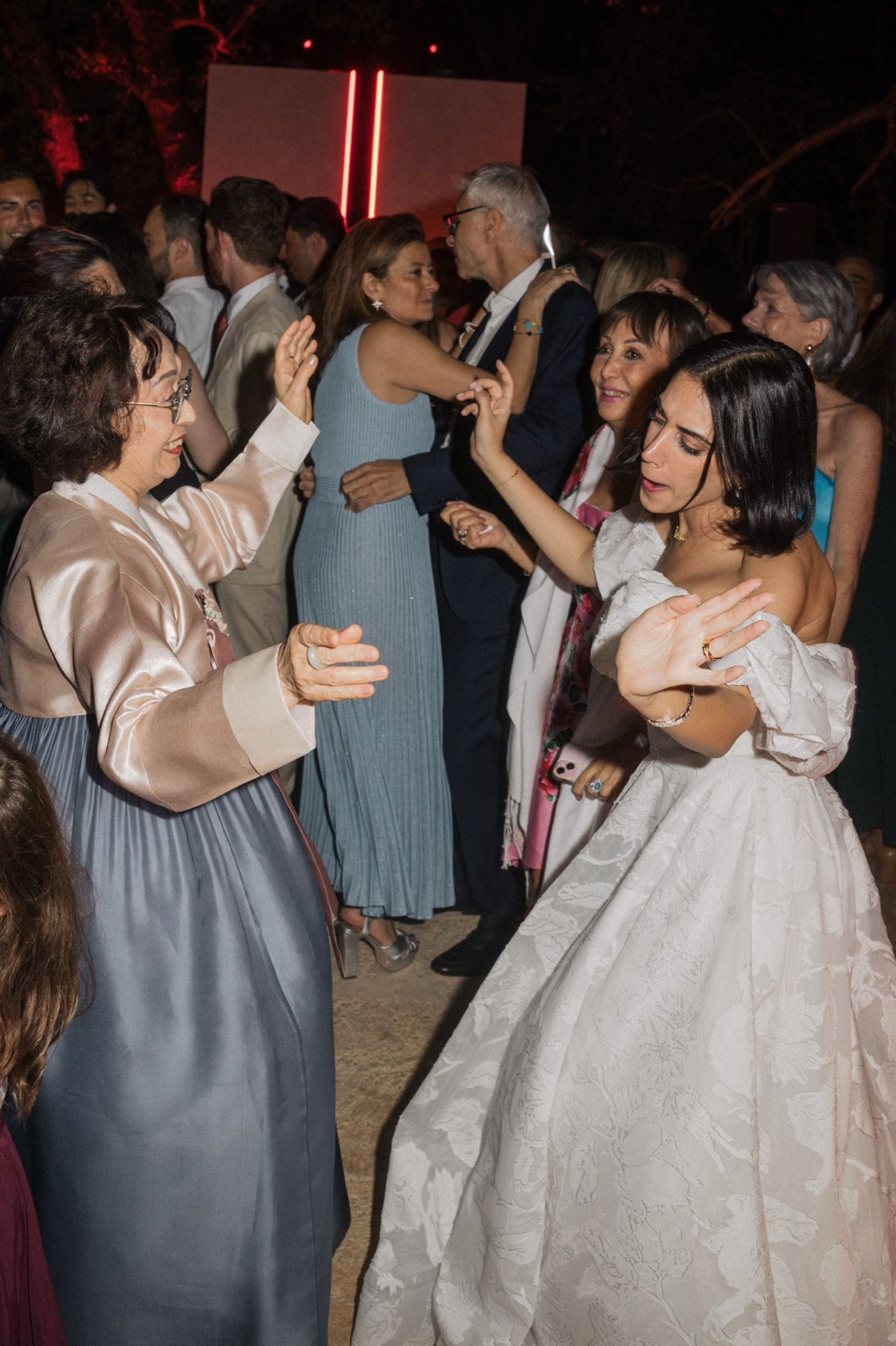 Guests dancing and celebrating at a wedding reception with dim lighting and neon red accents in the background.