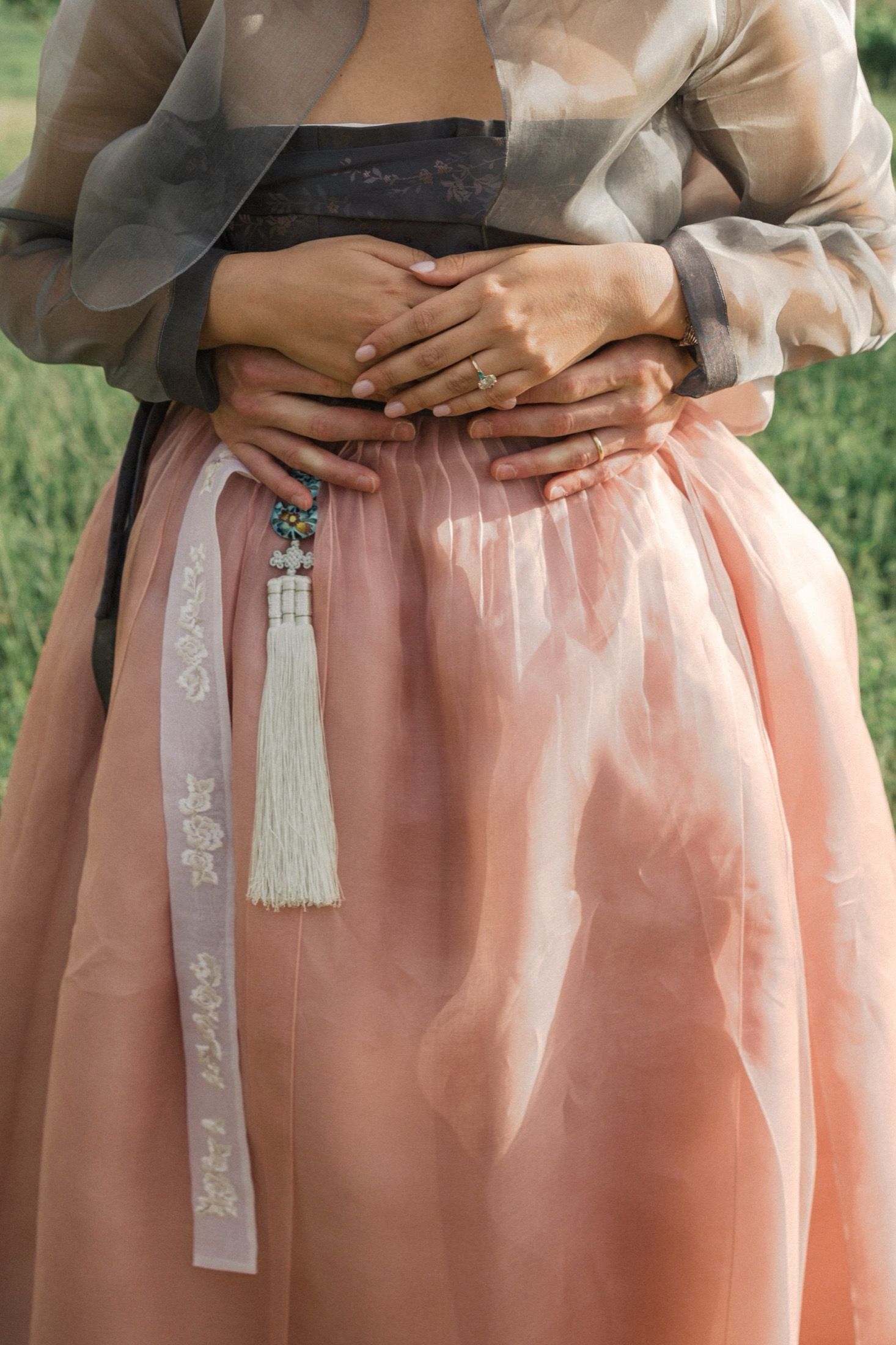 A person wearing a gray top and peach-colored hanbok skirt with white embroidered details and tassels.