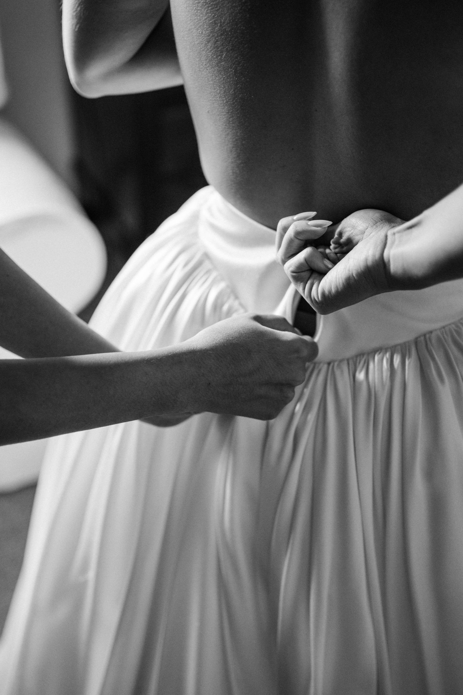 Black and white photo showing hands fastening buttons on the back of an elegant white wedding dress.