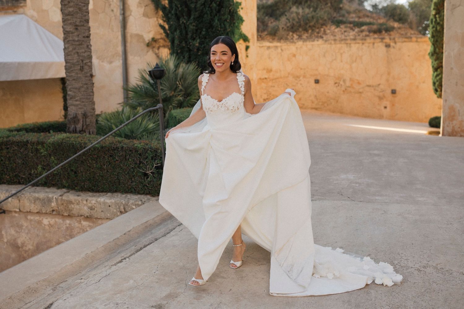 Bride in a flowing white wedding gown holding her dress train outdoors at a sunlit stone venue.