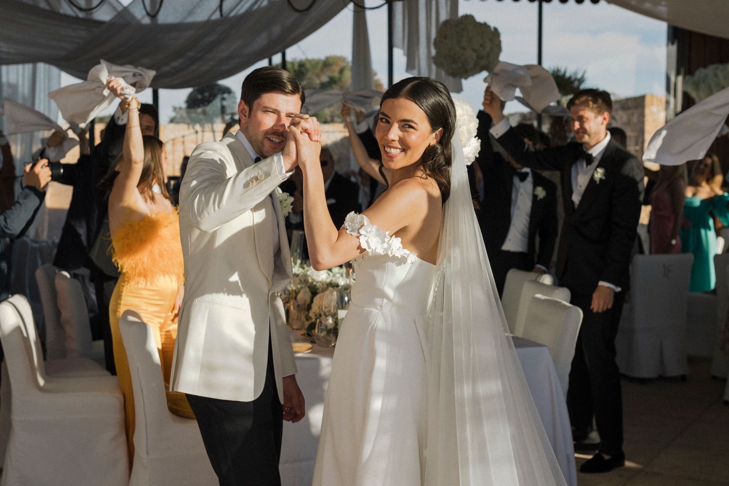 Bride and groom dancing joyfully at outdoor wedding reception, guests waving white handkerchiefs in celebration.