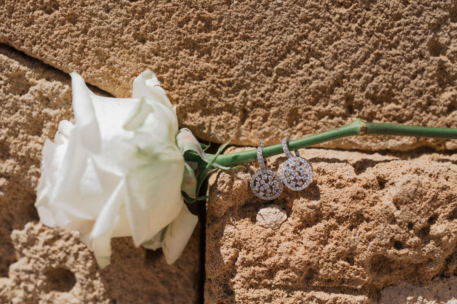 White rose and heart-shaped charm resting against a sandy textured stone wall in sunlight.