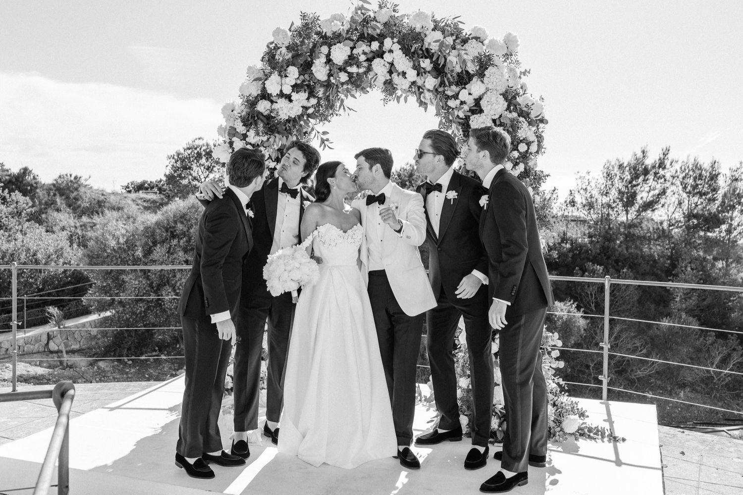 Black and white wedding photo of bride and groom with groomsmen under floral arch outdoors.