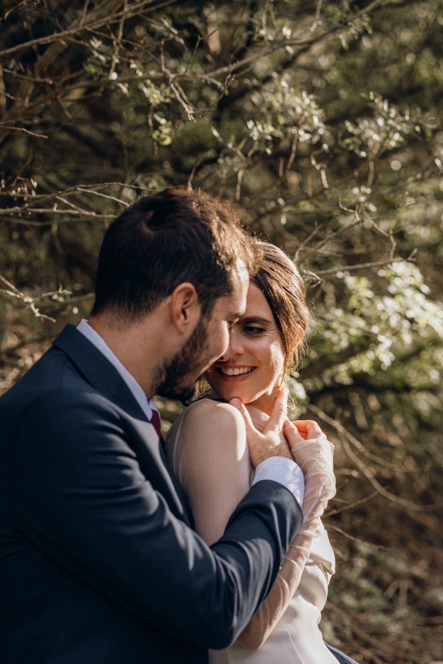 A romantic moment captured between a couple in formal wedding attire embracing under sunlit trees.