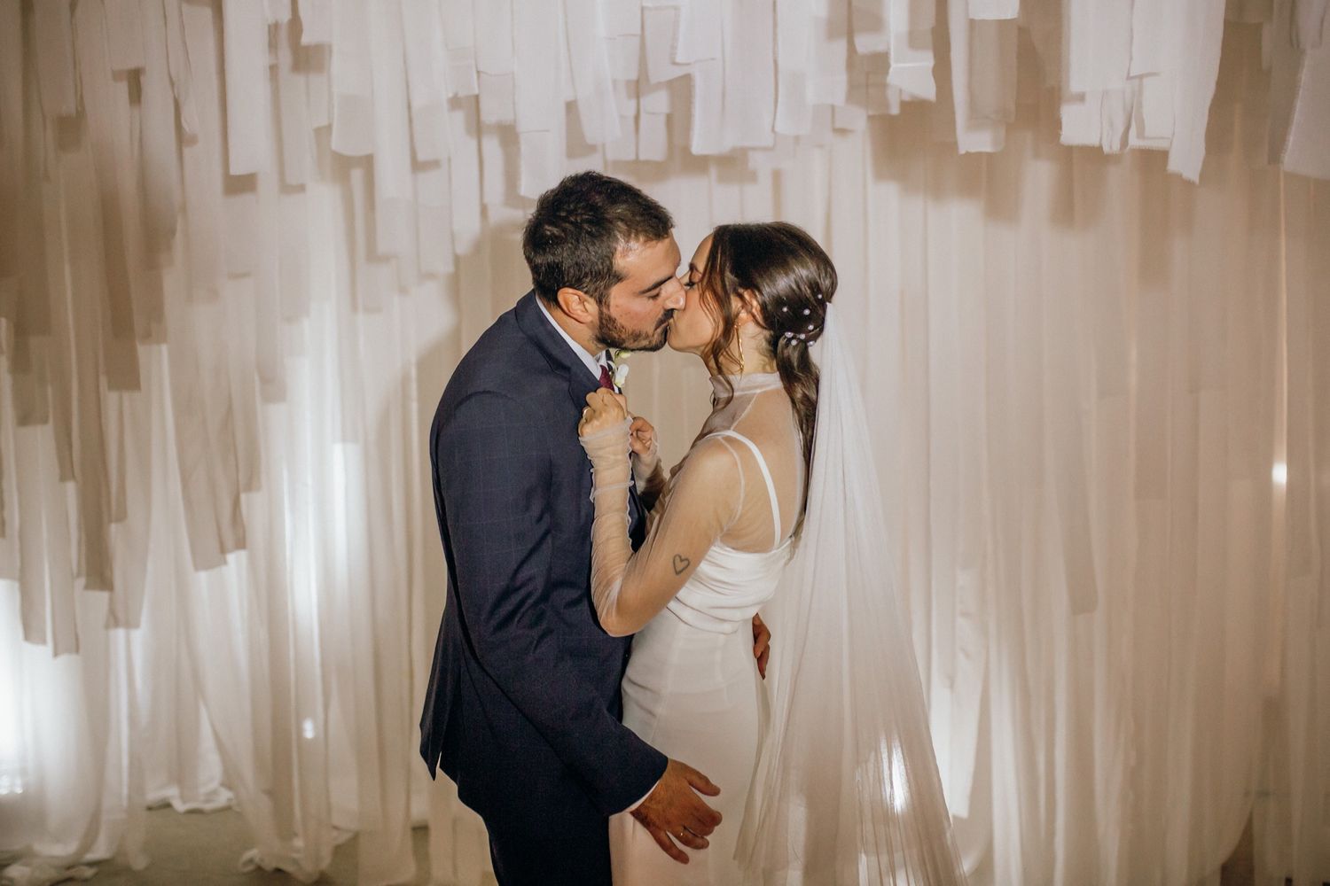 A romantic wedding couple shares a tender kiss in front of a wall decorated with flowing white fabric panels.