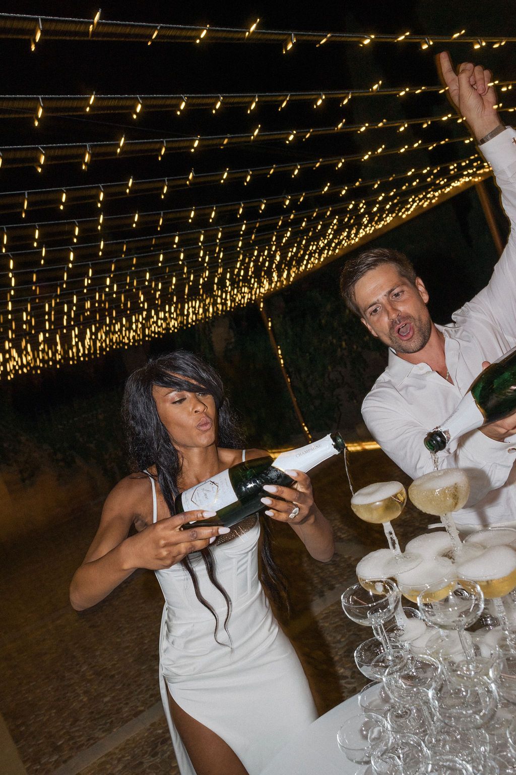 Wedding celebration under string lights with guests pouring champagne into stacked glasses.