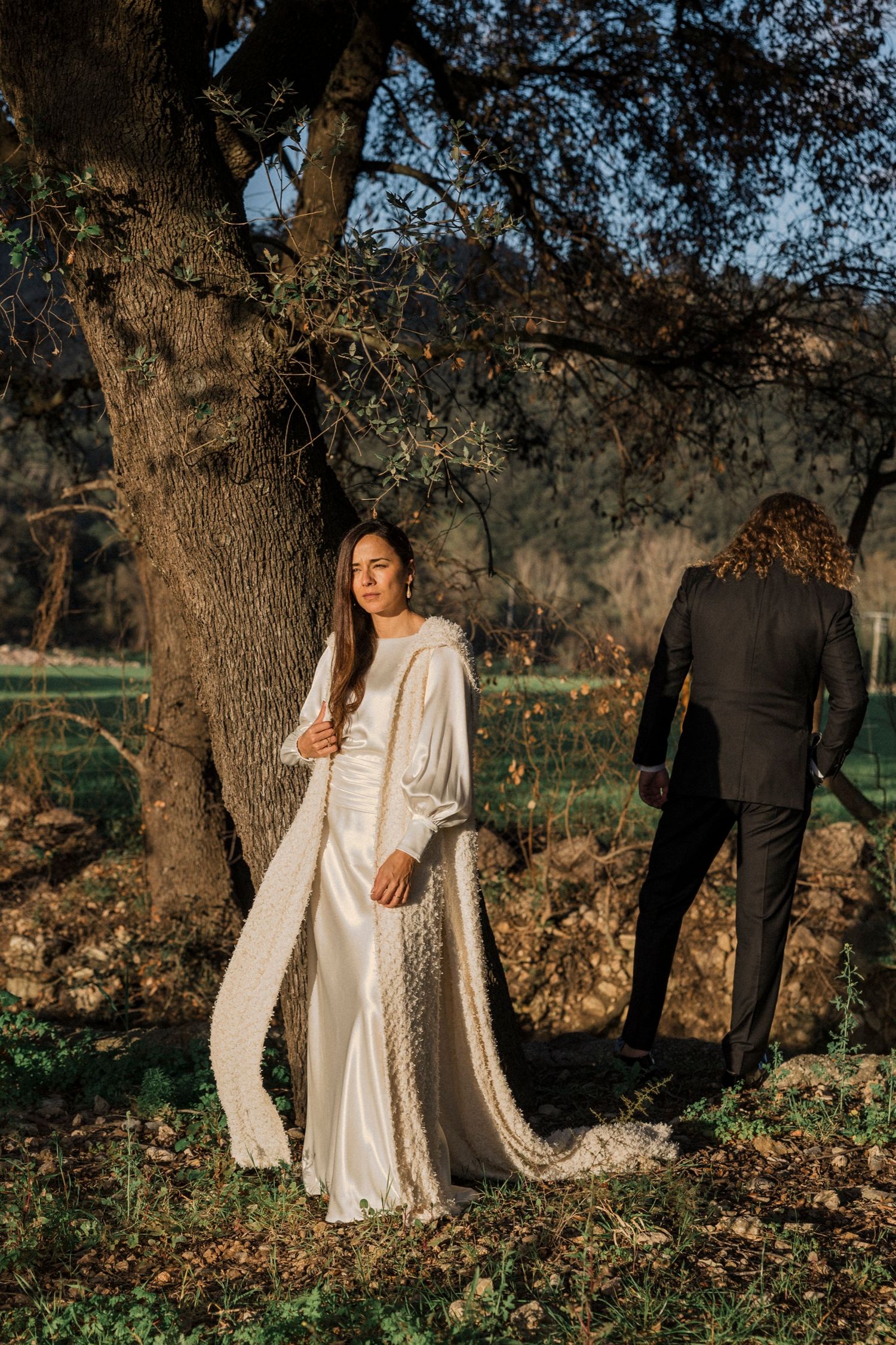 A couple in wedding attire poses romantically among trees during a sunset photoshoot.