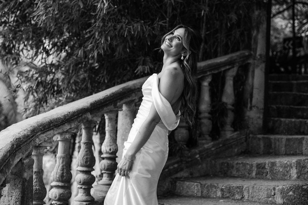 A bride in a white dress poses gracefully on stone steps with ornate balusters in a romantic garden setting.