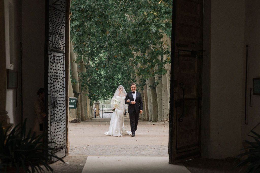 A wedding couple walks through an arched passageway framed by stone columns.