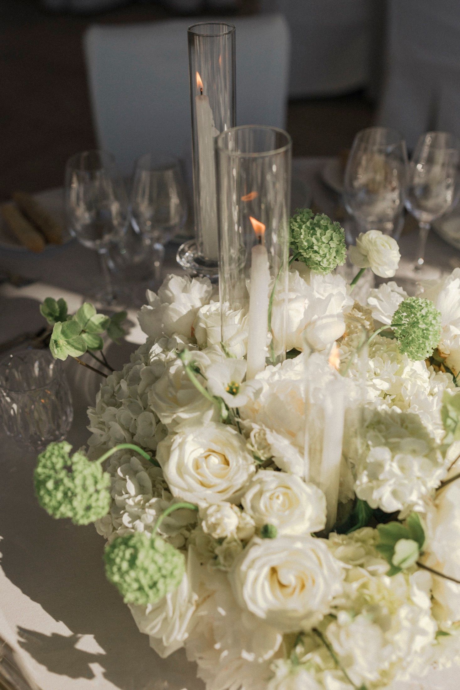 Elegant white floral centerpiece with roses, hydrangeas, and a lit candle in a glass hurricane vase on a wedding table.
