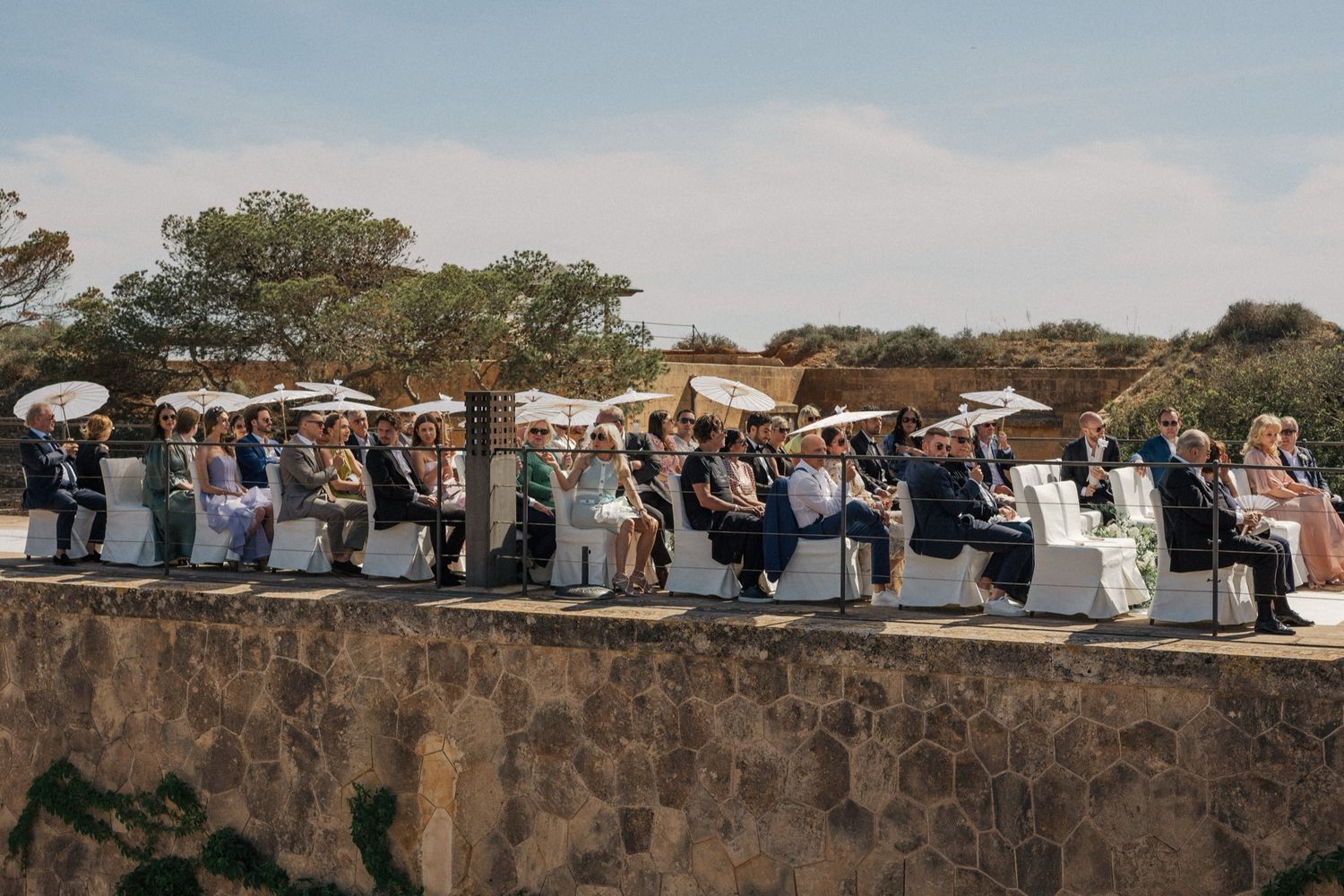 Outdoor wedding ceremony with guests seated in rows under white parasols on a stone terrace surrounded by natural landscape.