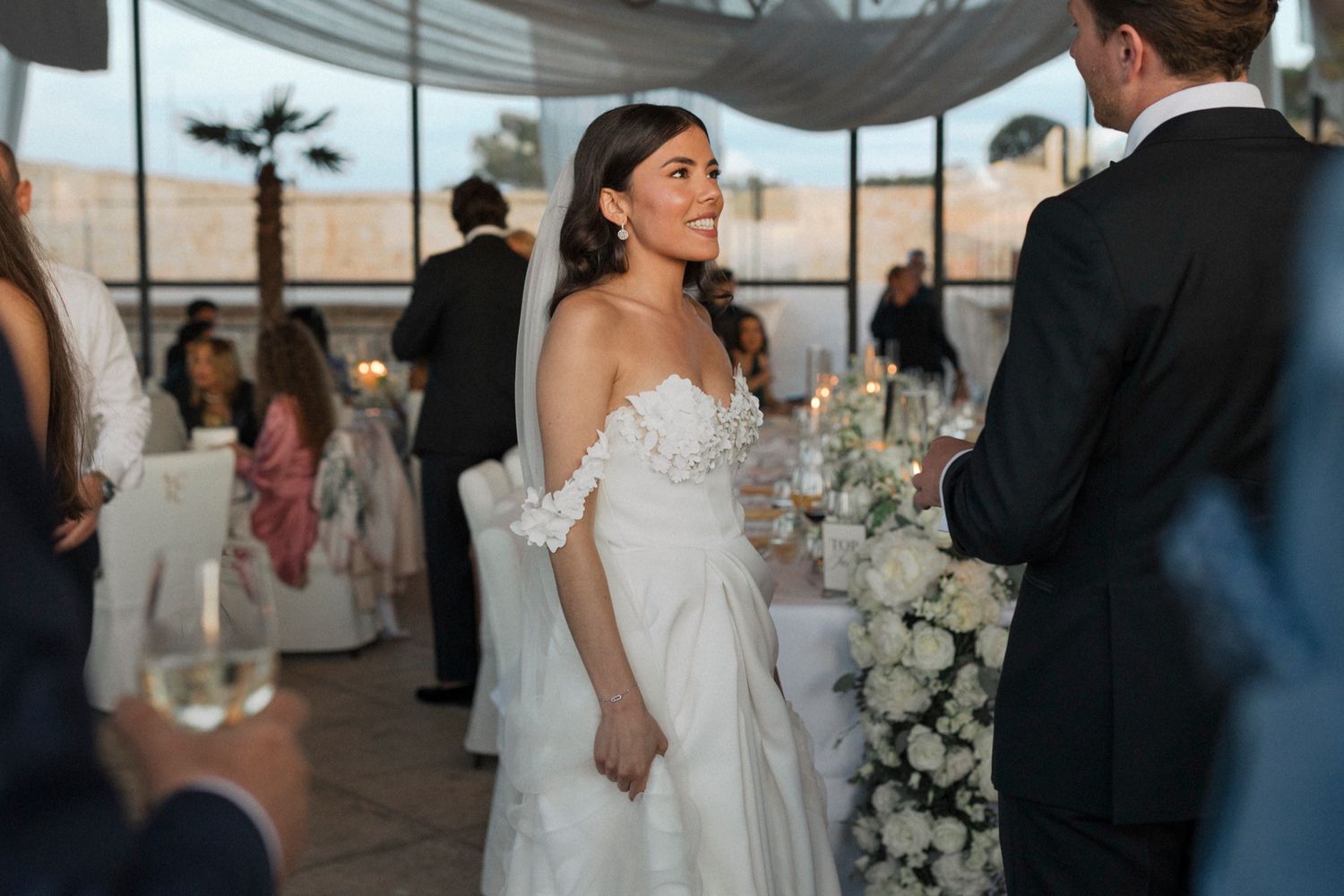 Bride in off-shoulder white gown smiling at wedding reception with floral arrangements and candles.