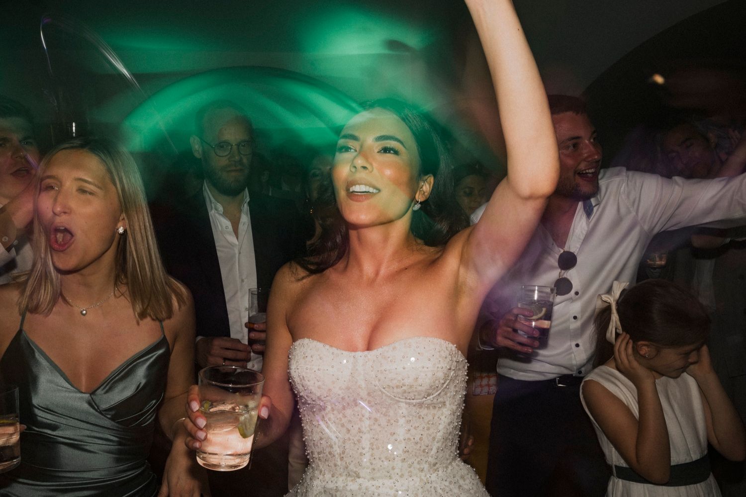 Bride in a sparkly strapless gown dancing joyfully at wedding reception with guests celebrating around her.
