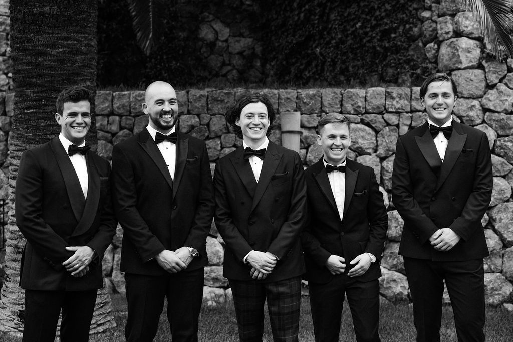 Groomsmen in black tuxedos lined up against stone wall for formal wedding photo.