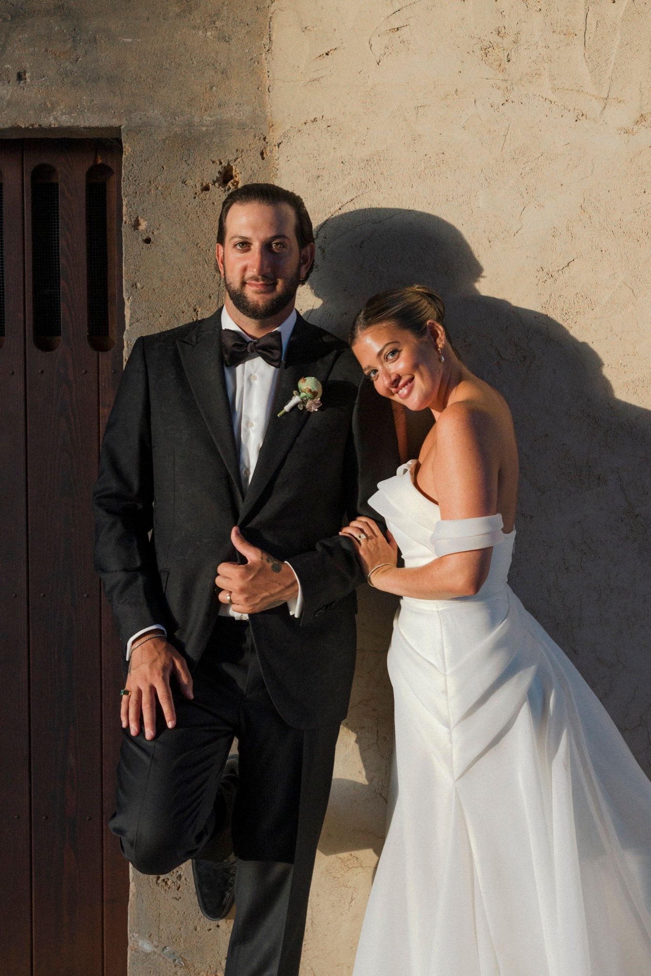 A joyful wedding portrait of a couple posing against a rustic wall with dramatic shadows.