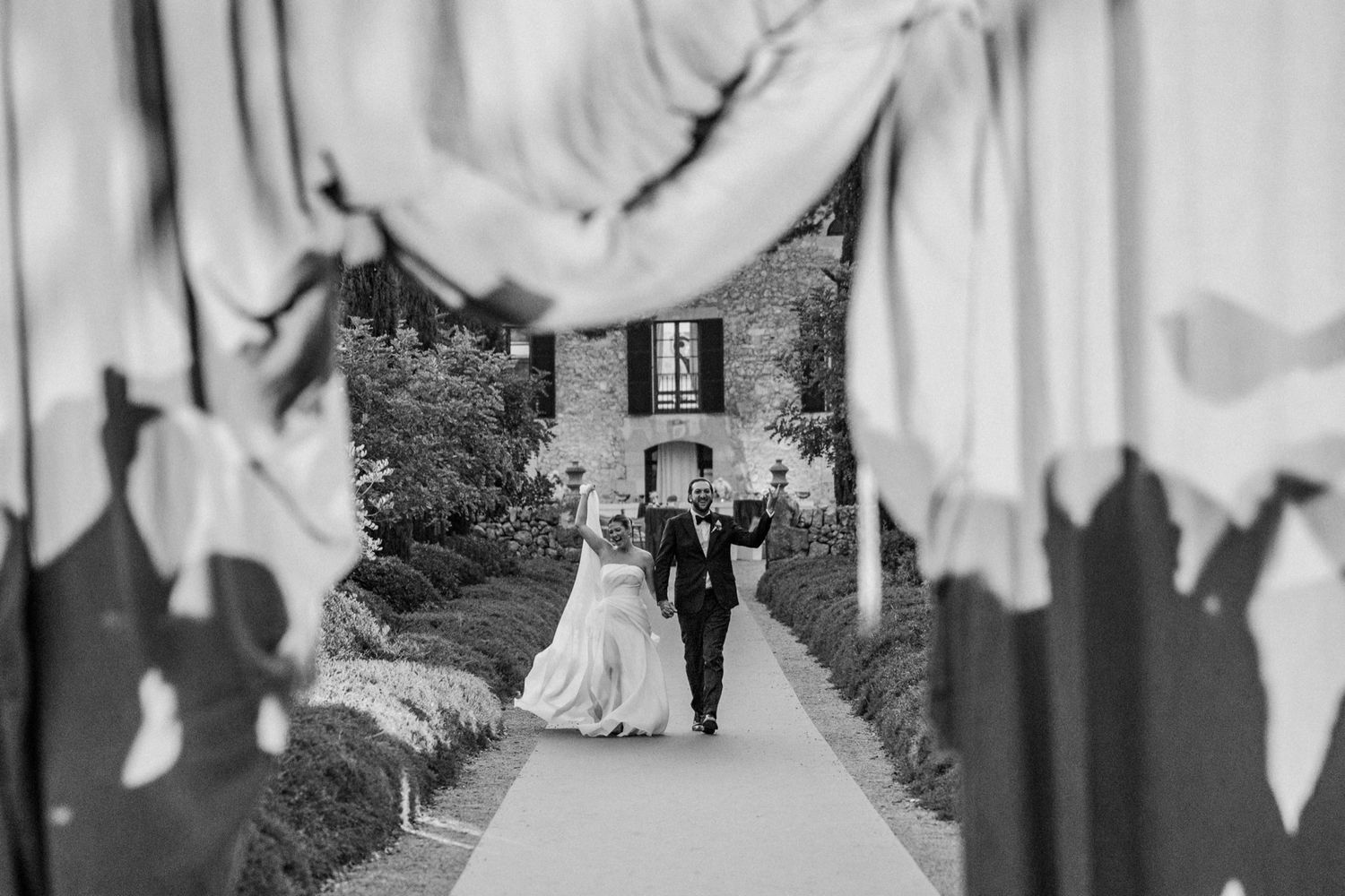 Black and white wedding photo framed by guest silhouettes as couple walks down path.