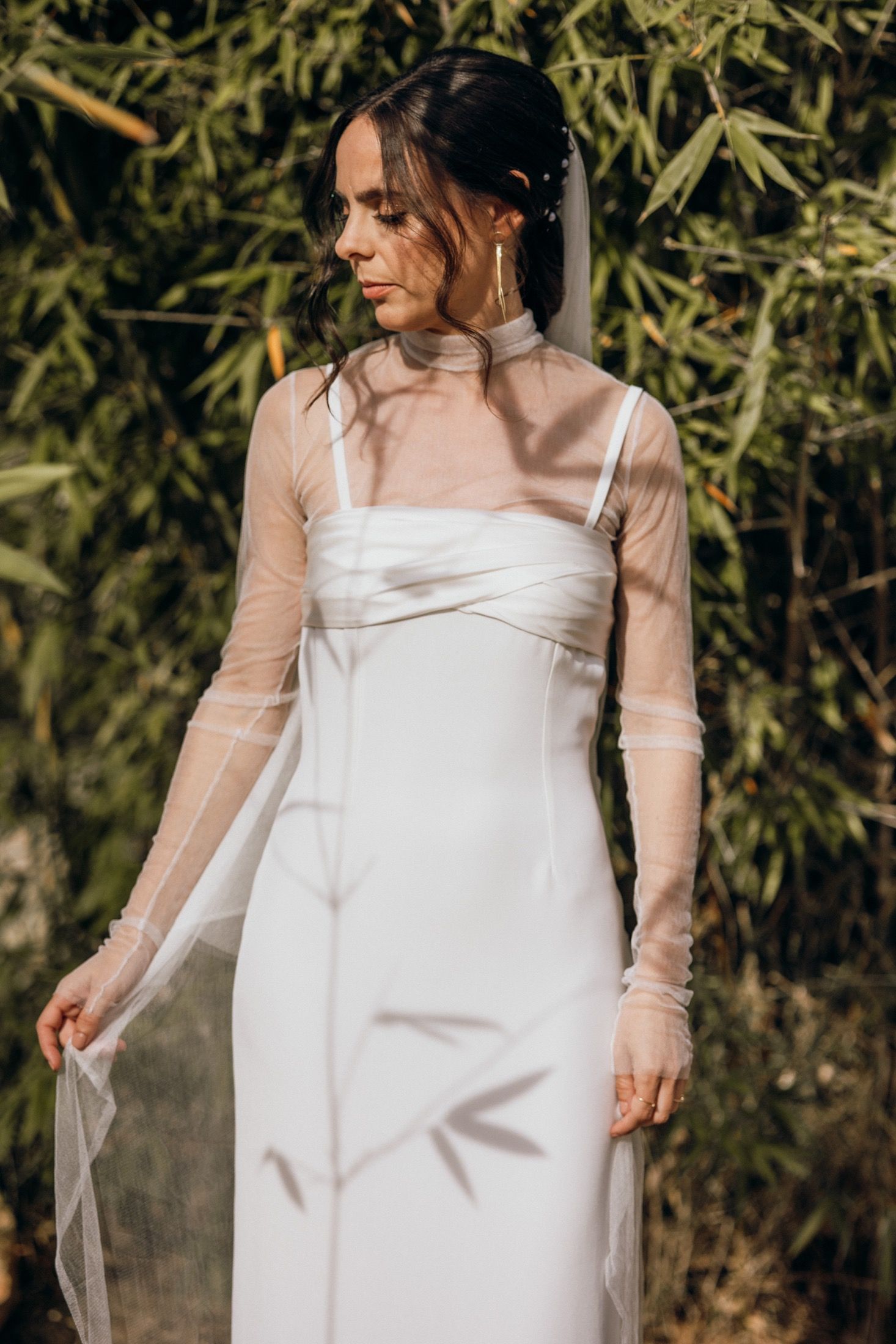 A person in a white wedding dress with sheer sleeves poses against a natural bamboo backdrop in warm sunlight.