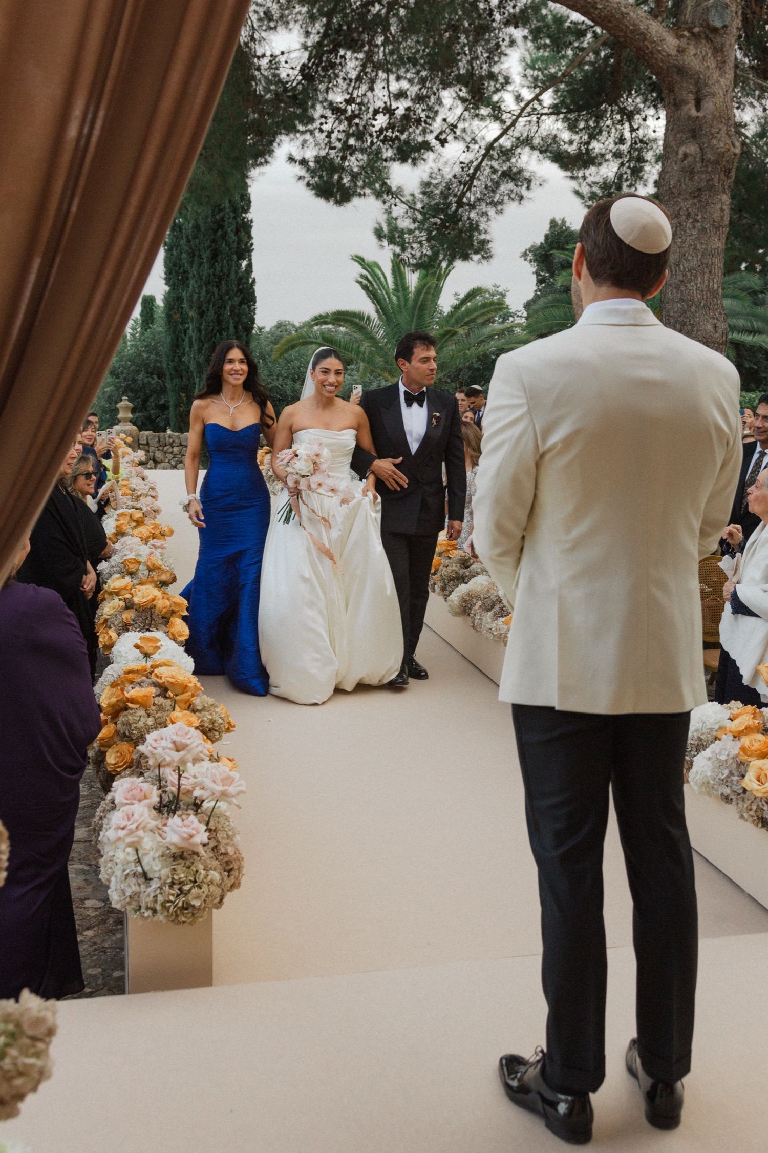 Wedding ceremony guests gather outdoors on a terrace with floral arrangements and palm trees in the background.