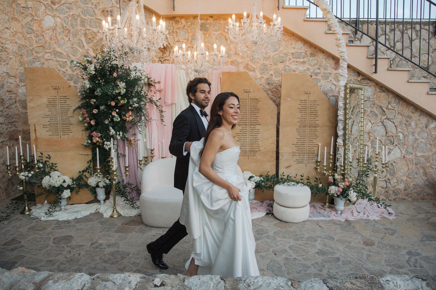 A newlywed couple celebrates under a crystal chandelier with floral decorations and stone wall backdrop.