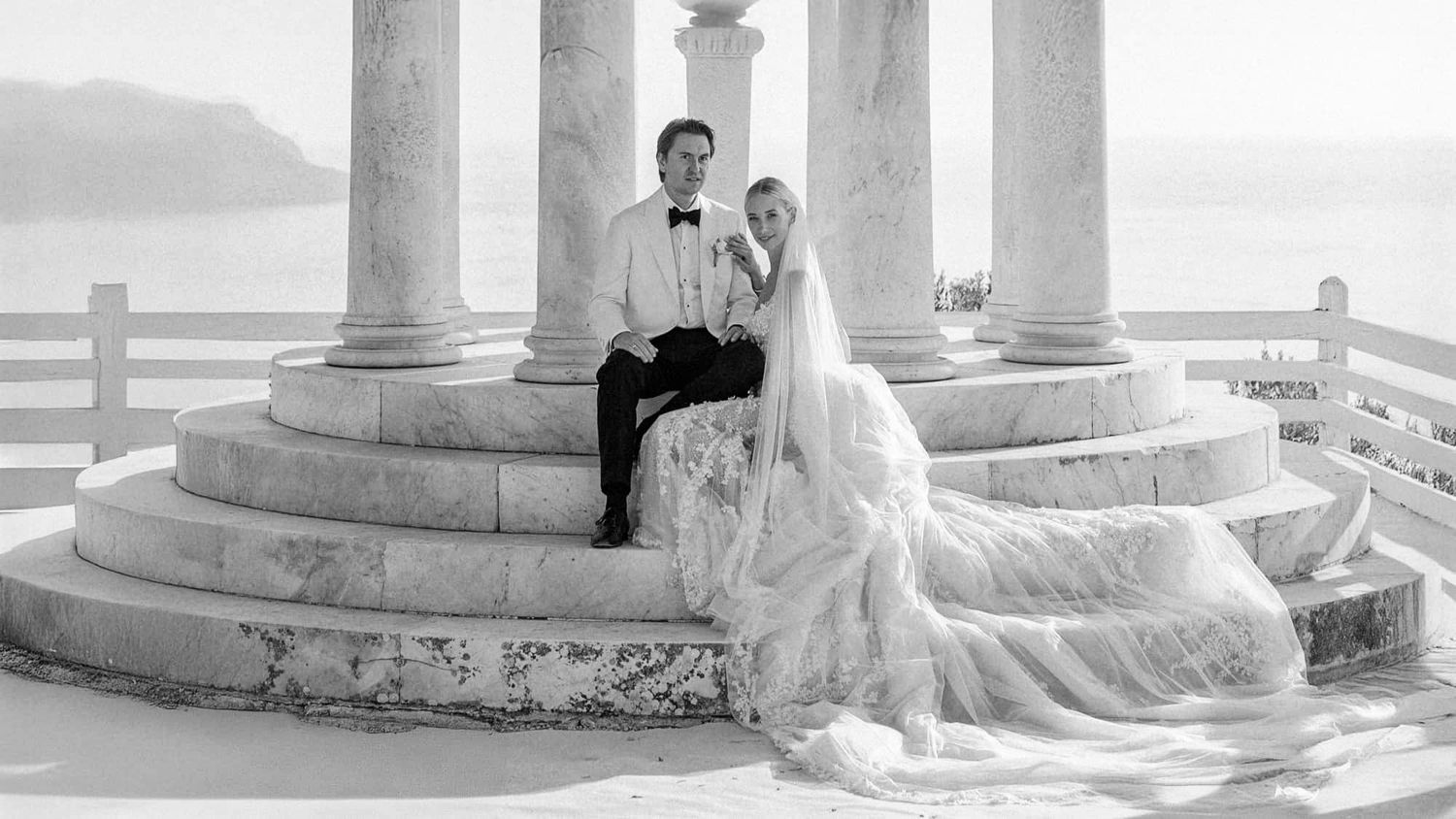 A romantic black and white wedding photo of a couple sitting on elegant stone steps with white columns.