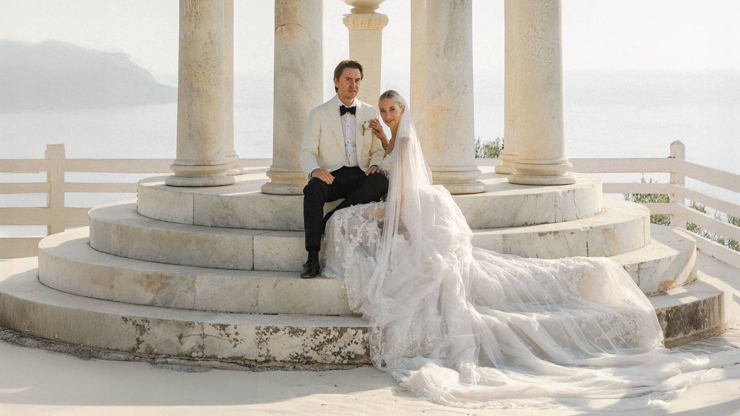 A couple sits on elegant white stone steps with ornate columns overlooking a misty coastal view. Son Marroig