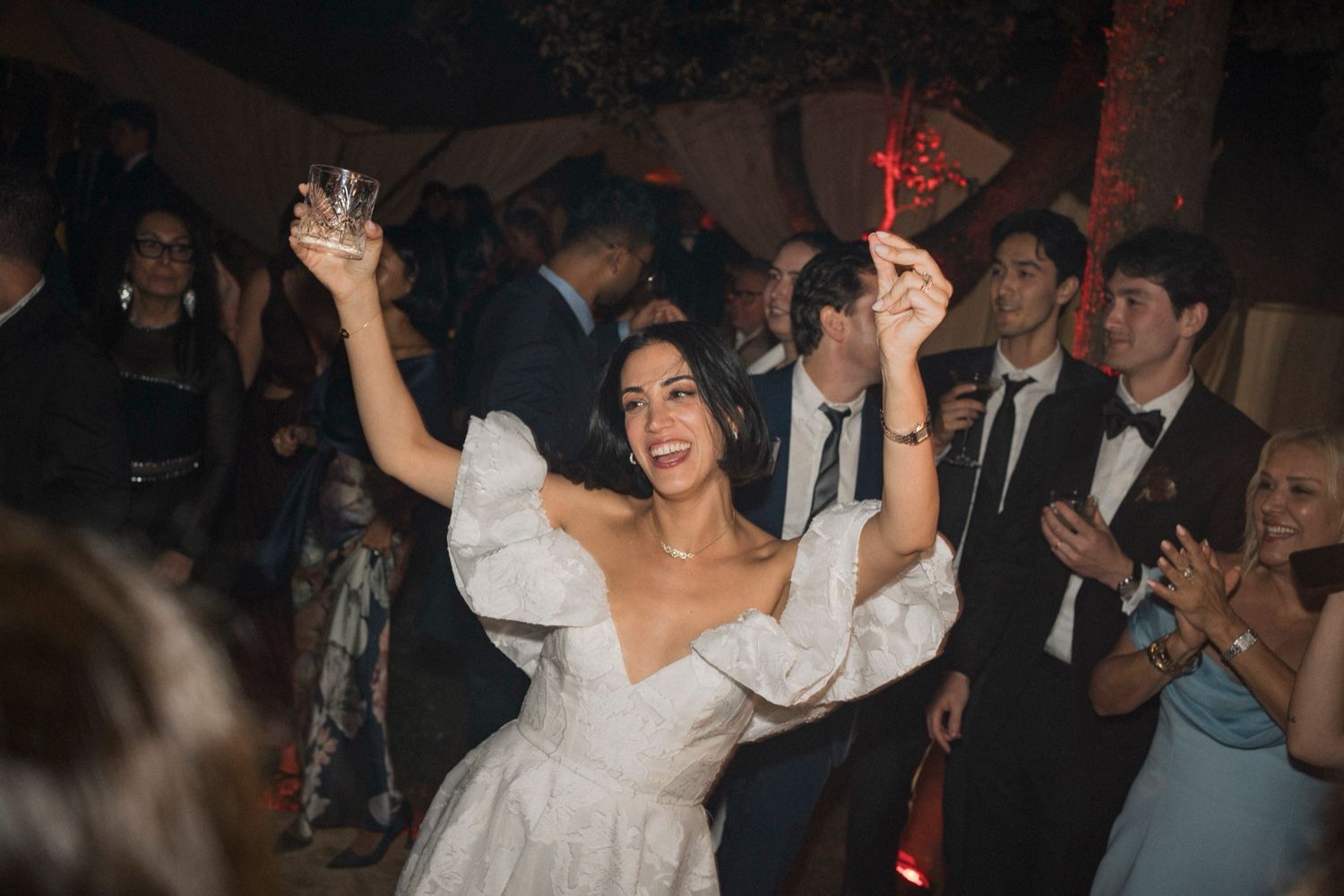A person in a white wedding dress dances joyfully with raised arms on a dark dance floor surrounded by party guests.