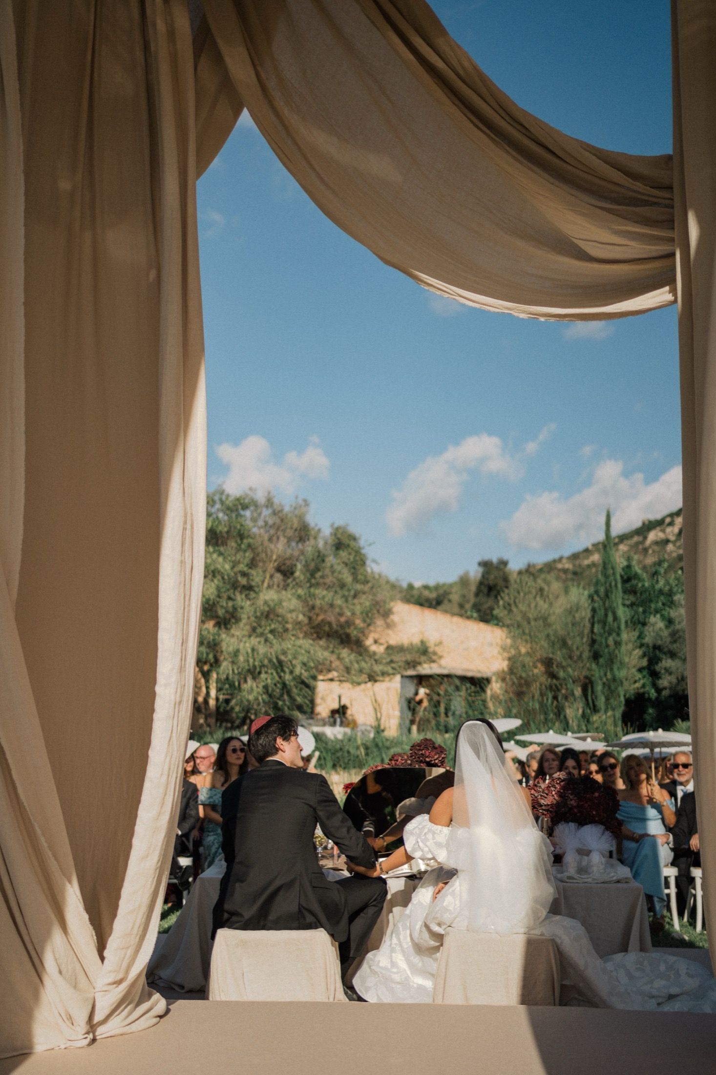 A wedding ceremony under draped beige fabric with guests seated outdoors against a scenic hillside backdrop.