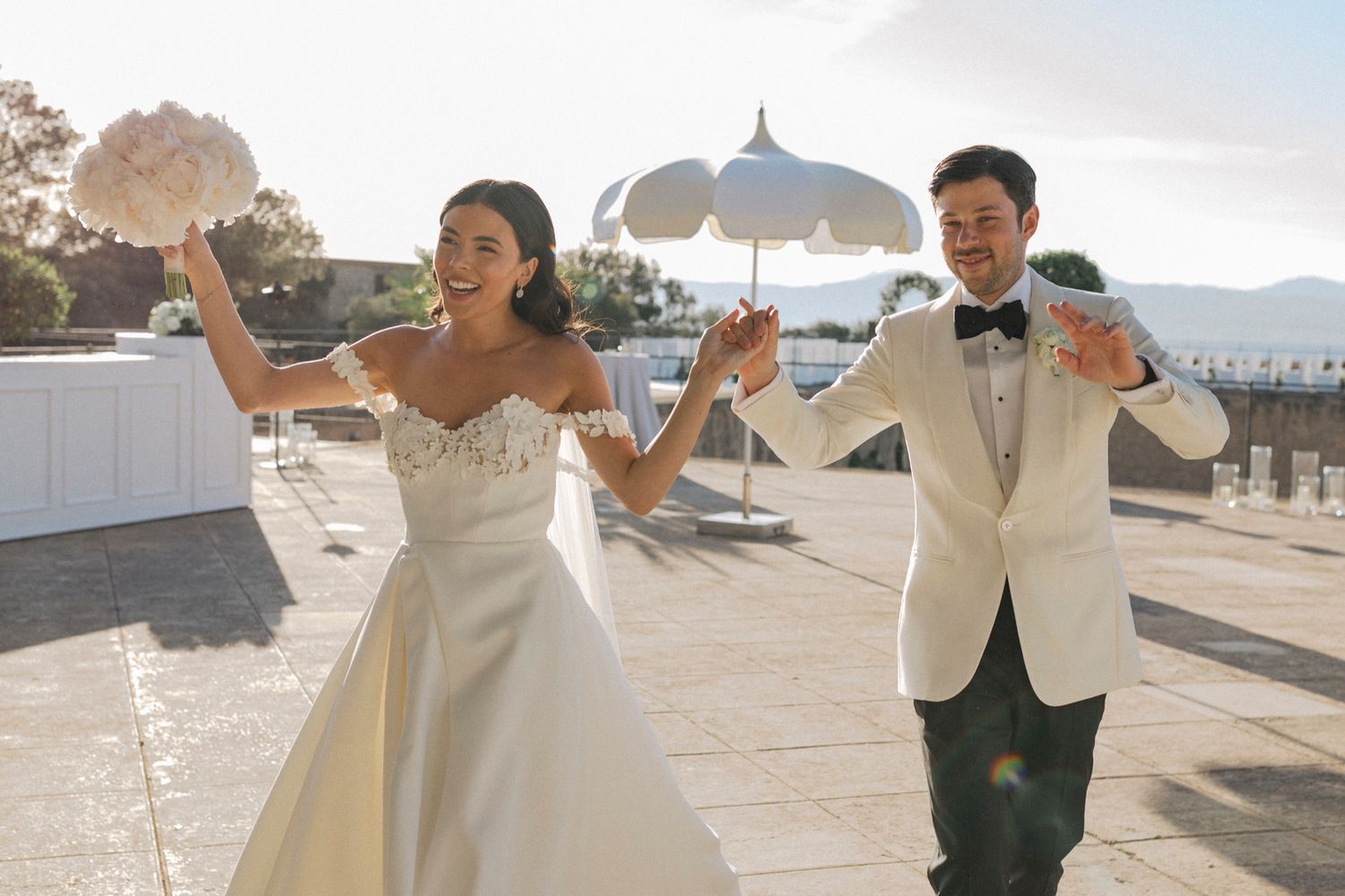 Joyful bride and groom celebrating outdoors on sunny terrace, bride holding bouquet aloft in elegant white gown and tuxedo.
