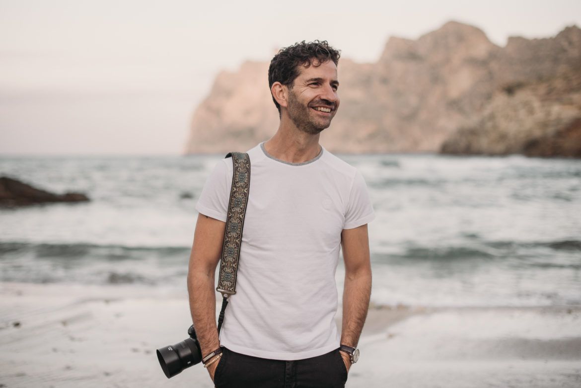 A person in a white t-shirt stands on a rocky beach with ocean waves and cliffs in the background.