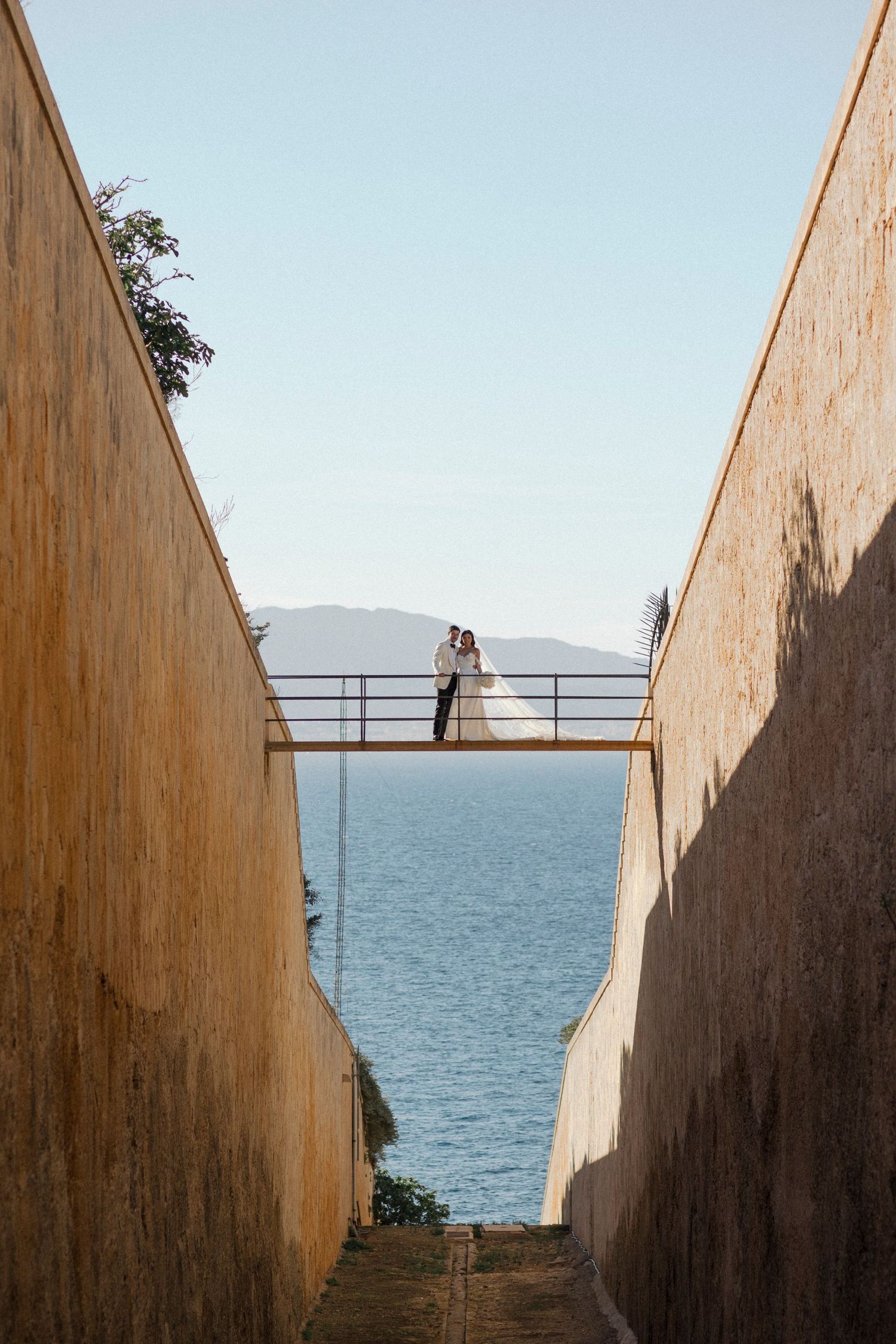 Bride and groom embracing on a bridge between stone walls overlooking the sea and mountains.