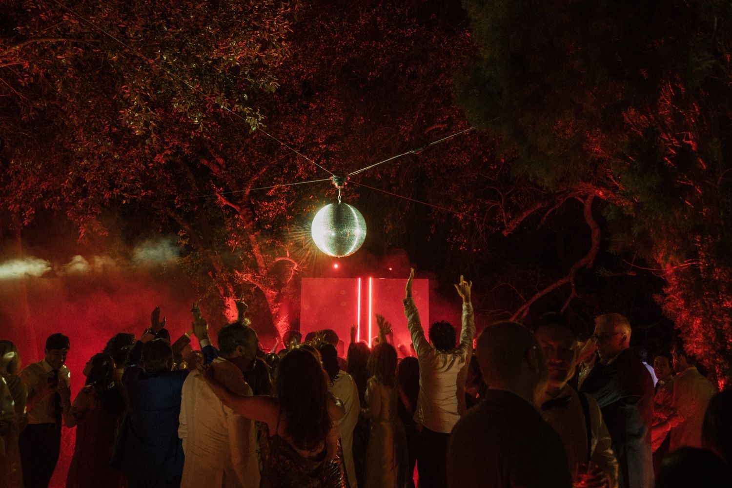 Silhouettes of people gather under trees at night with glowing red lights creating an eerie atmospheric scene.