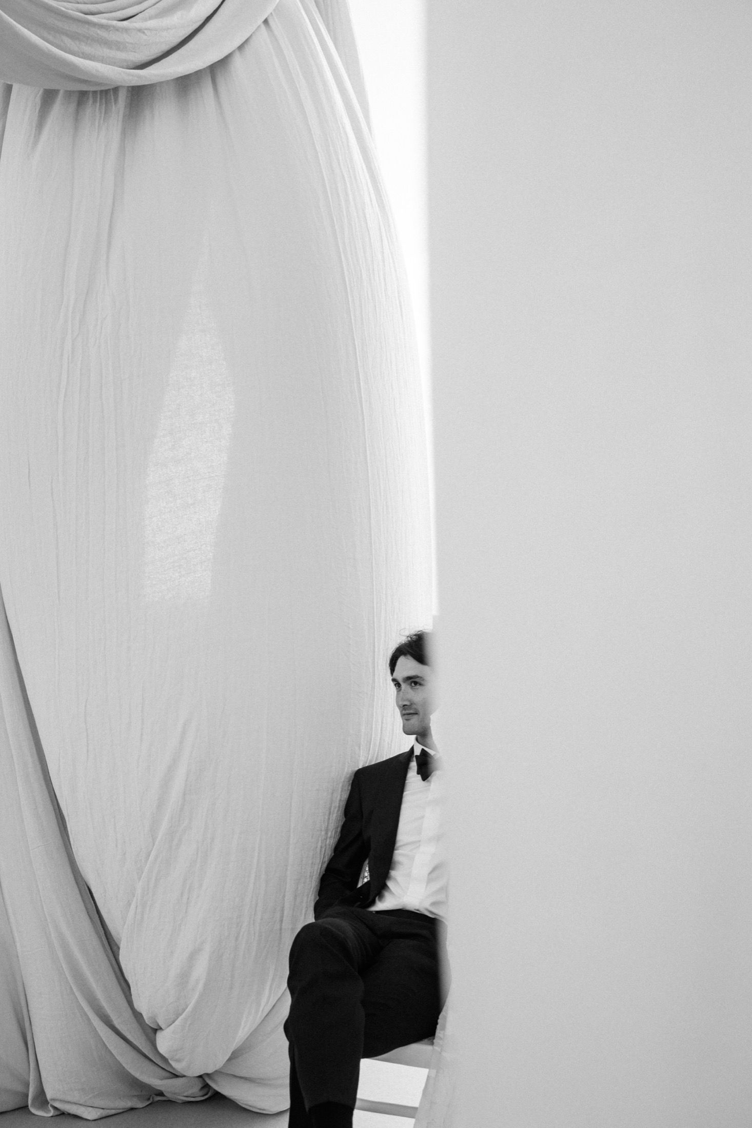 A black and white artistic portrait showing someone peeking from behind flowing white curtains in formal attire.