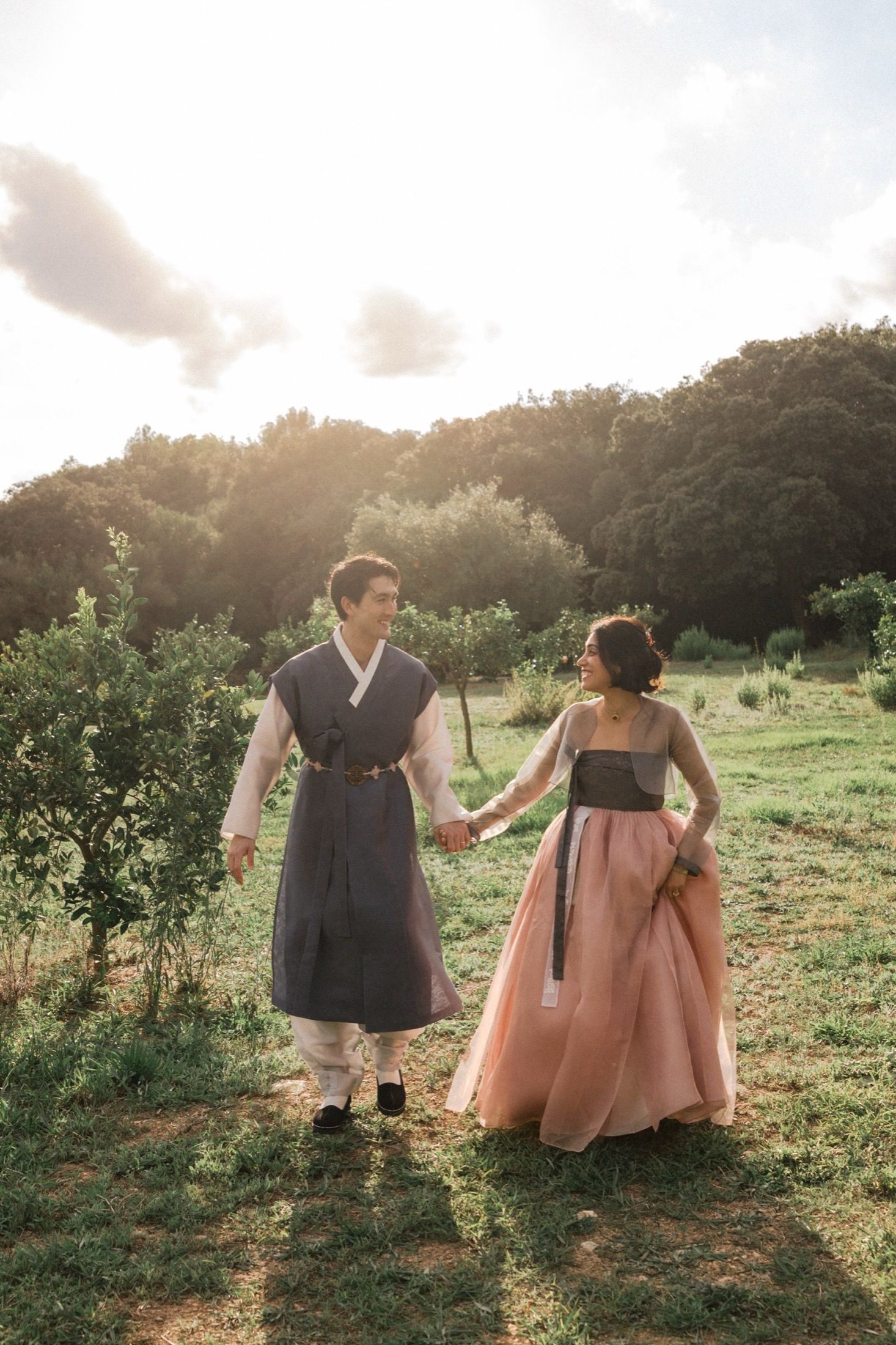 Two people in traditional hanbok dresses walk hand in hand through a sunlit garden at golden hour.