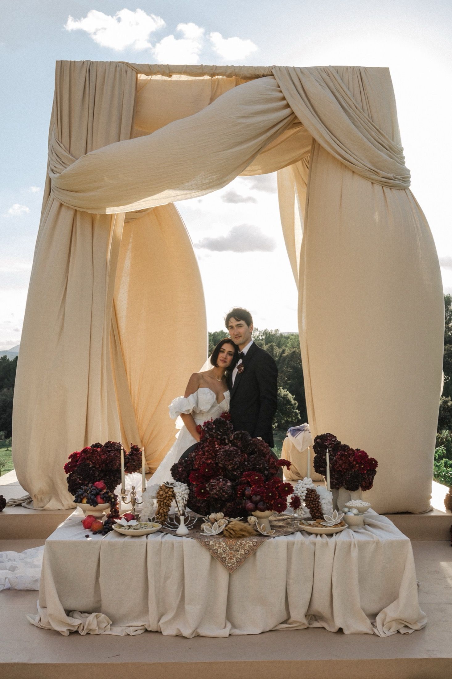 Elegant outdoor wedding reception setup with white drapes, burgundy floral arrangements and formal table settings.