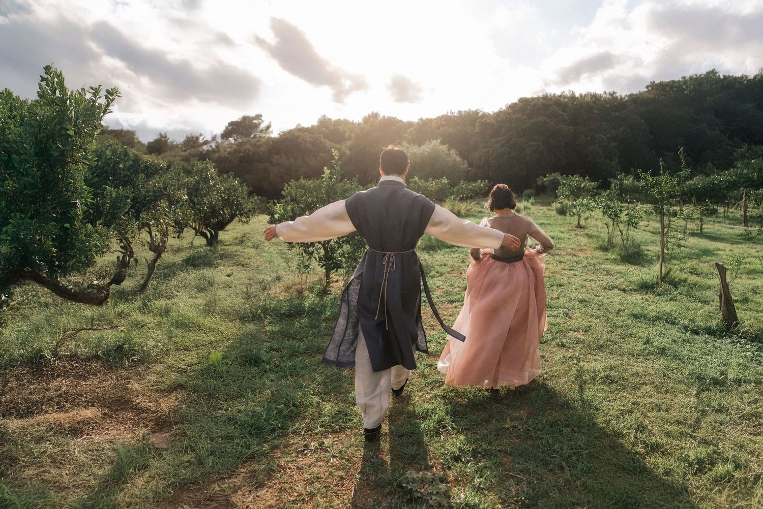 Couple in formal attire walks through a sunlit meadow during a golden hour photoshoot.