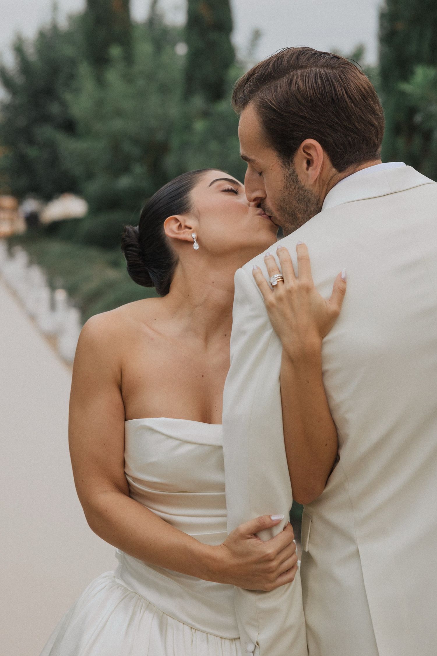A romantic wedding couple sharing a tender kiss outdoors, with the bride in white and groom in cream attire.
