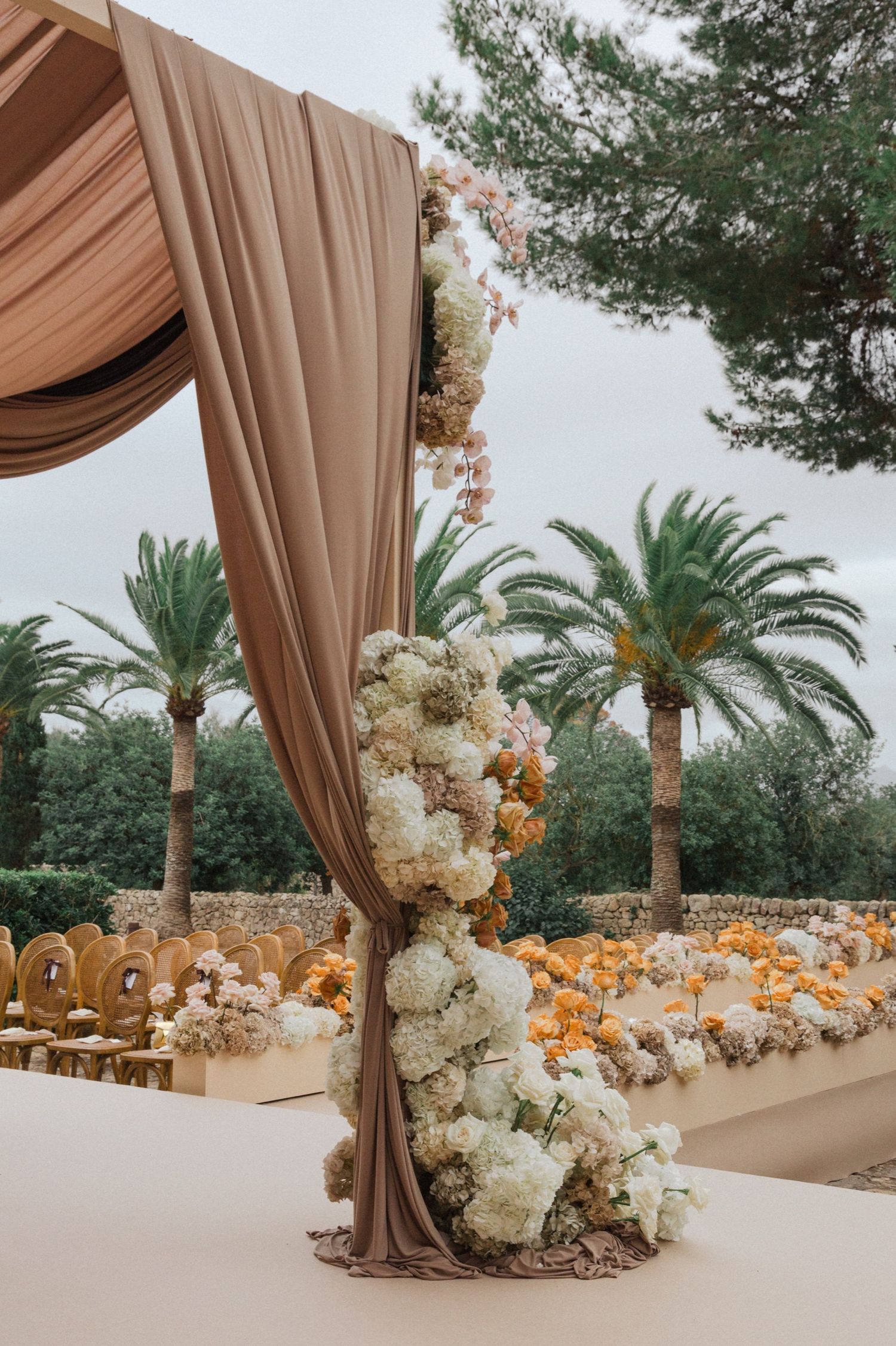 Elegant outdoor wedding ceremony setup with draped brown fabric and white floral arrangements against palm trees.