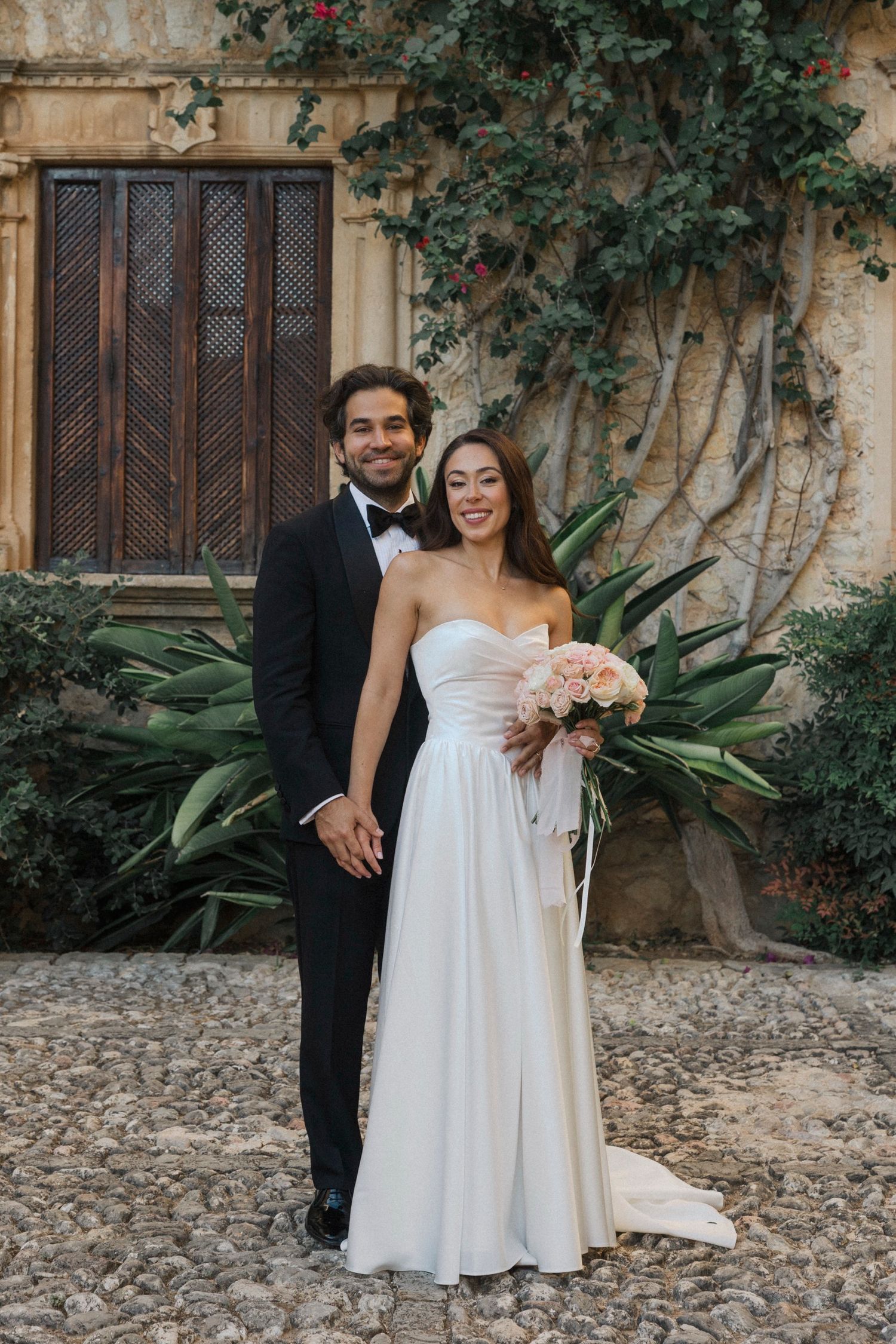 A couple poses together on their wedding day in front of a rustic stone wall with climbing vines and wooden door.