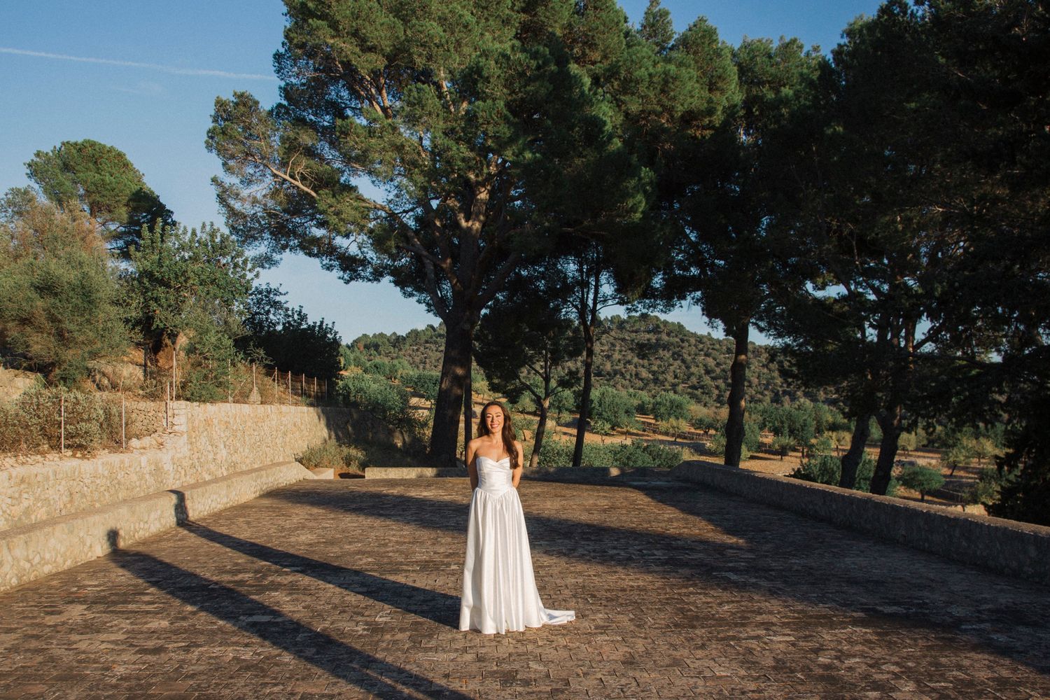 A person in a white dress walks through a scenic path lined with tall pine trees and golden sunlight.