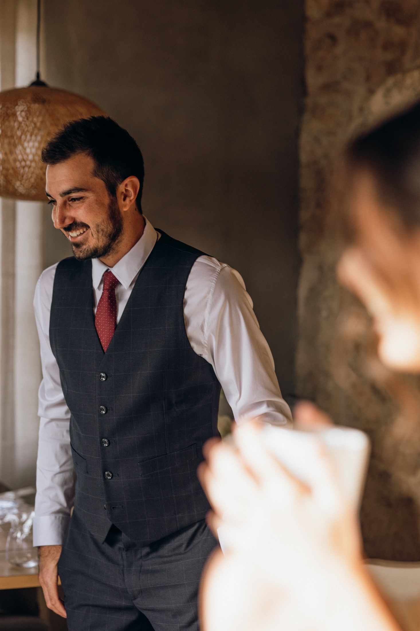 A person wearing a dark vest and white shirt smiles warmly while getting dressed in a rustic indoor setting.