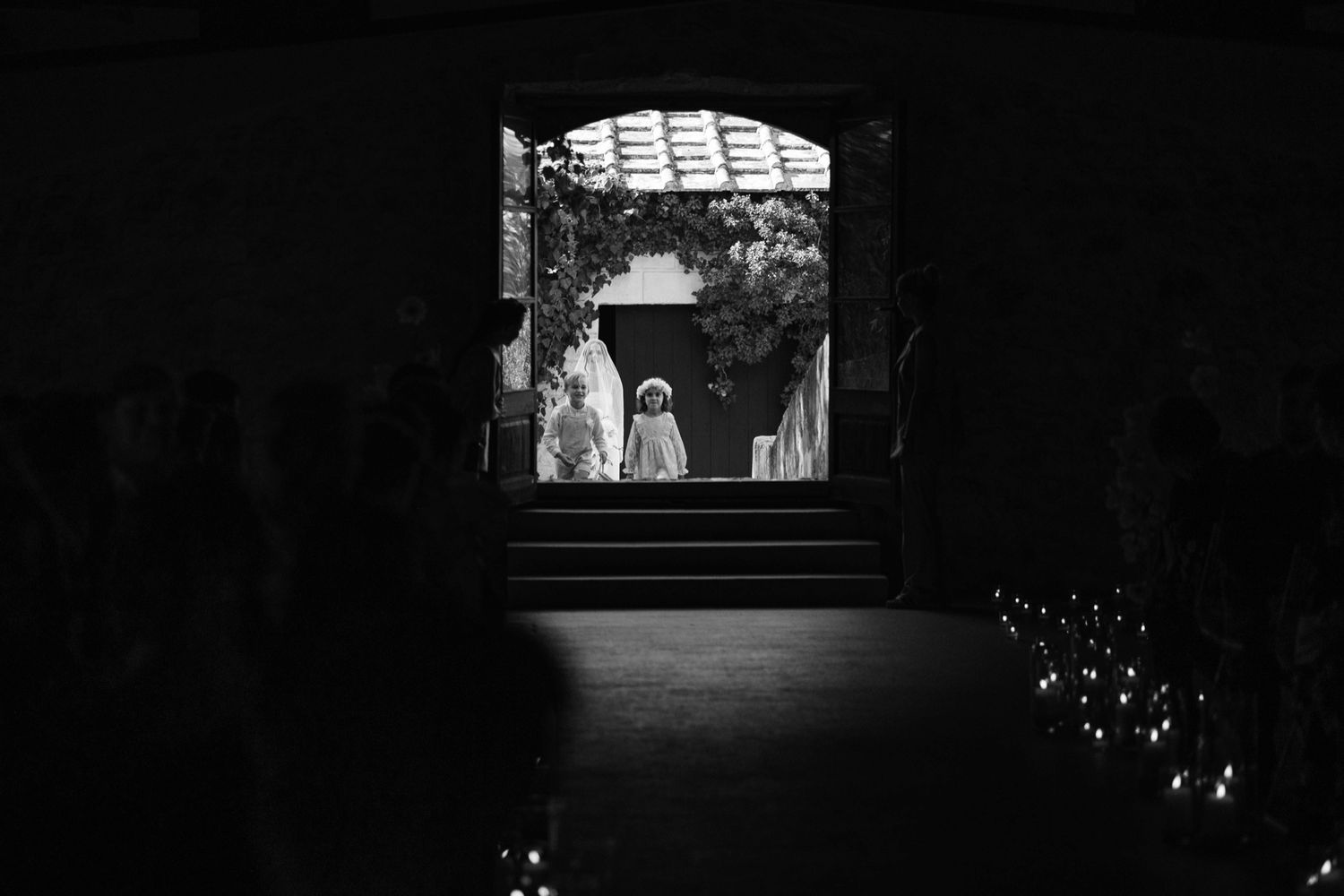 Black and white photo of a wedding ceremony viewed through an archway with glowing candles lining the path.