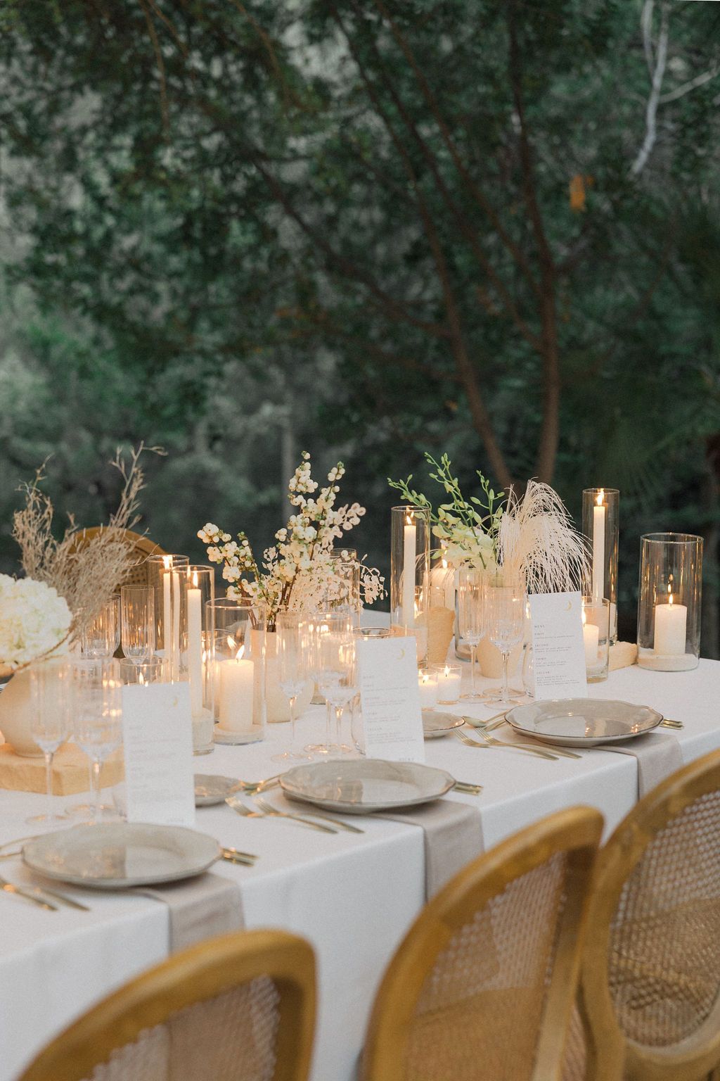 Romantic outdoor wedding table setting with candles and white linens under trees at dusk.
