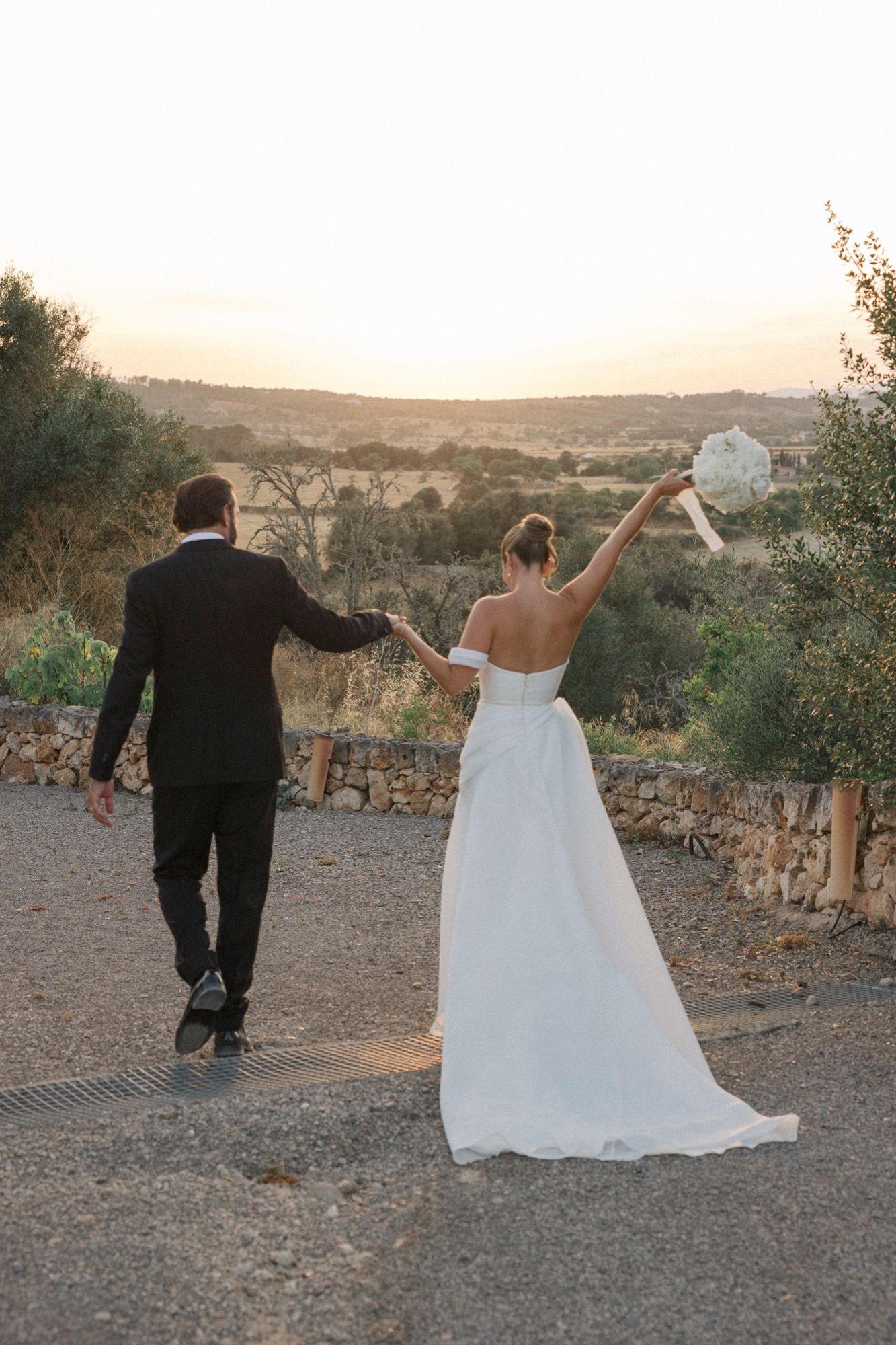 Wedding couple celebrates with raised arms while walking through scenic outdoor setting at sunset.