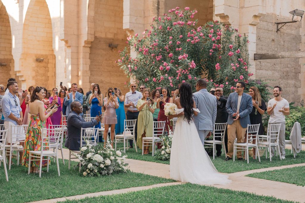 Wedding guests seated in white chairs within a historic stone courtyard surrounded by blooming flowers.