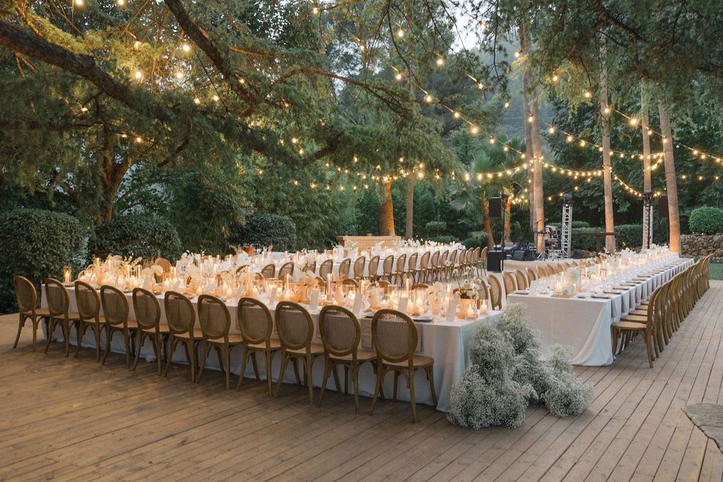 Long banquet tables with white linens set for wedding reception under string lights and trees.