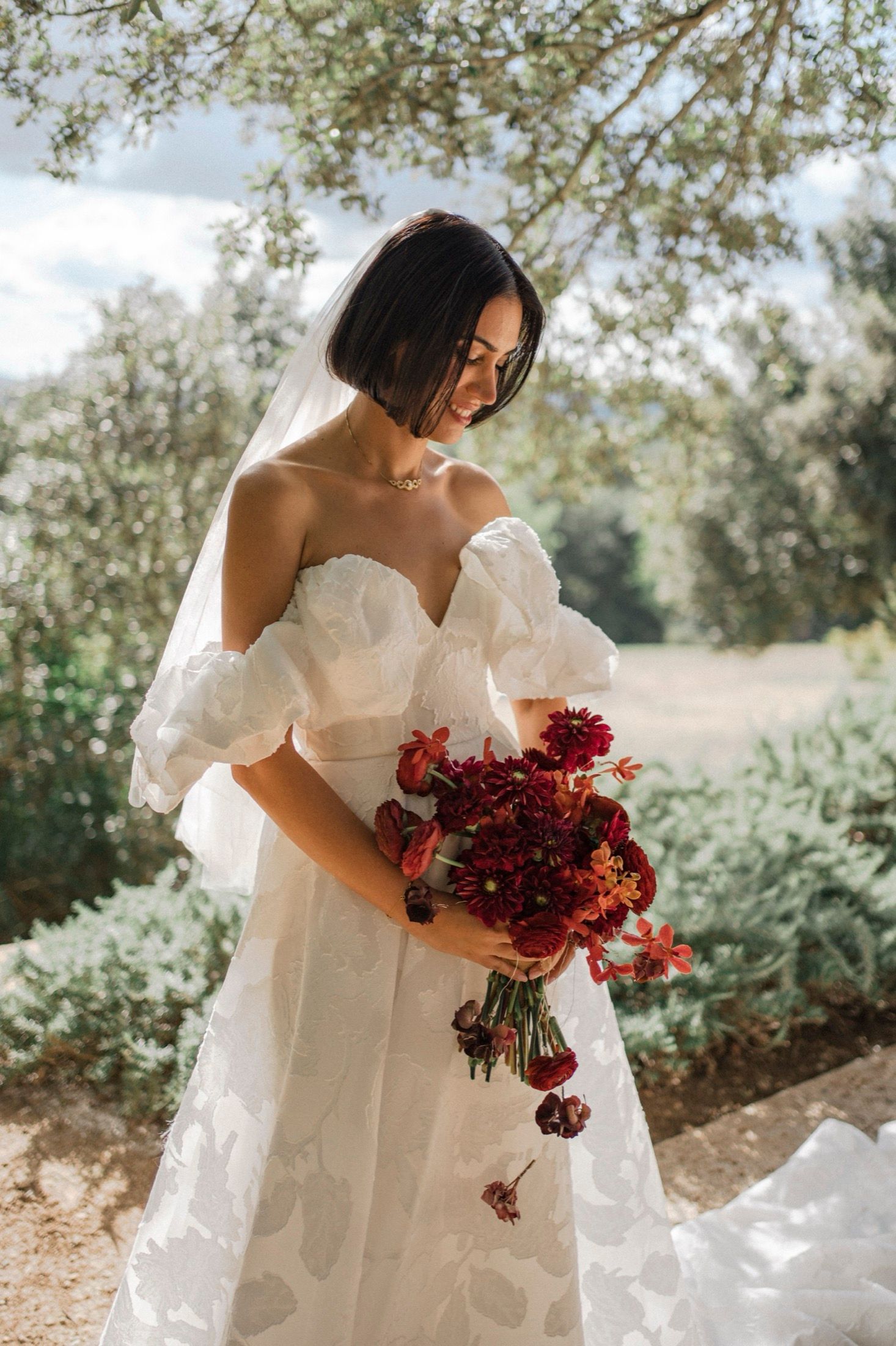 Bride in white wedding dress holds vibrant red and burgundy floral bouquet under sunlit trees in garden setting.