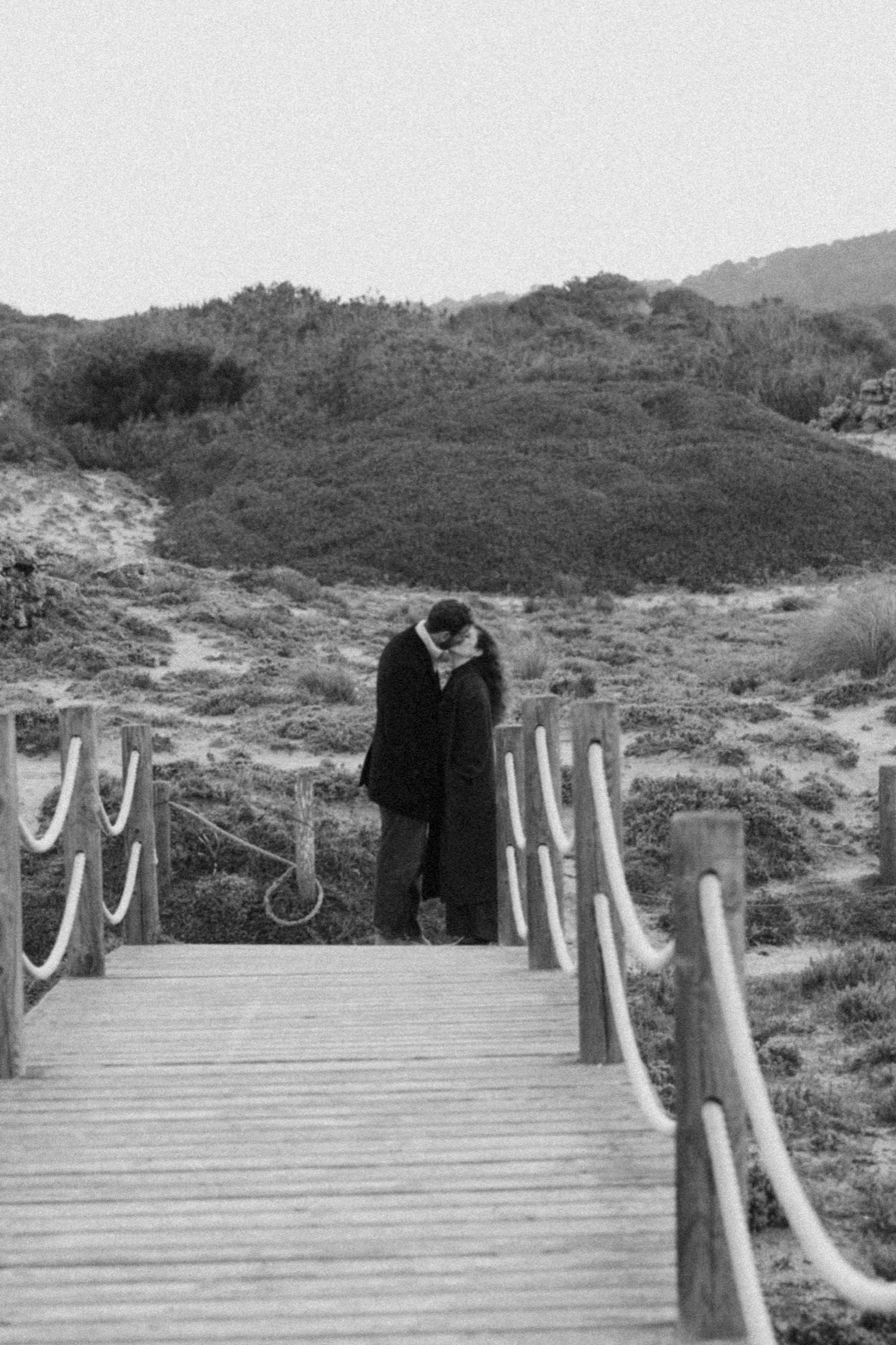 Black and white photo of a wooden boardwalk path through coastal sand dunes with two figures in the distance.
