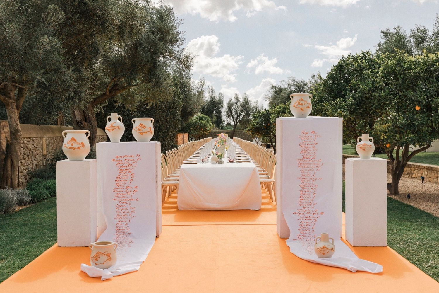 Elegant outdoor wedding ceremony setup with white pedestals and orange carpet runner leading to altar in garden setting.