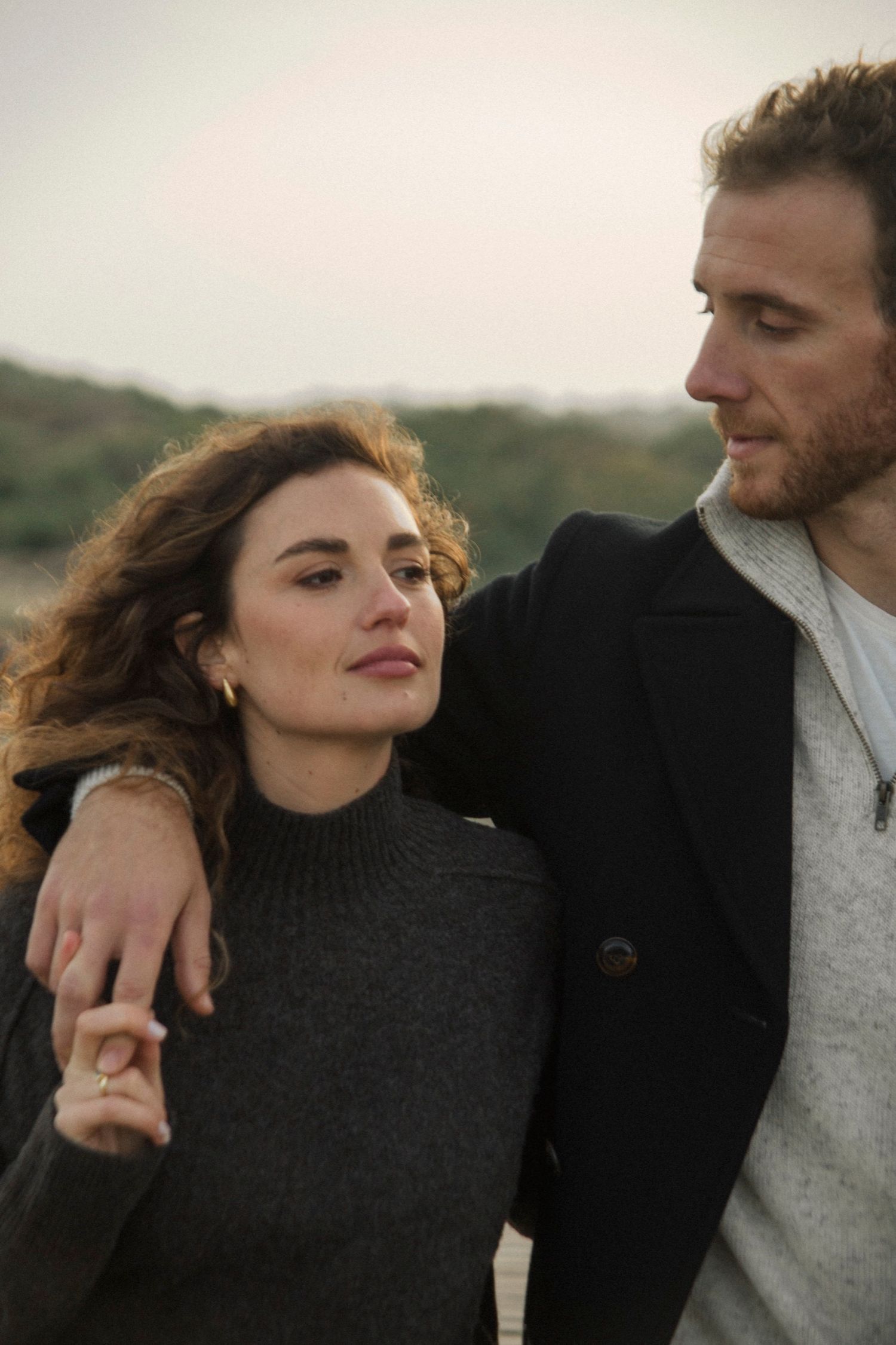 A romantic couple embraces outdoors while wearing dark sweaters against a soft natural backdrop.